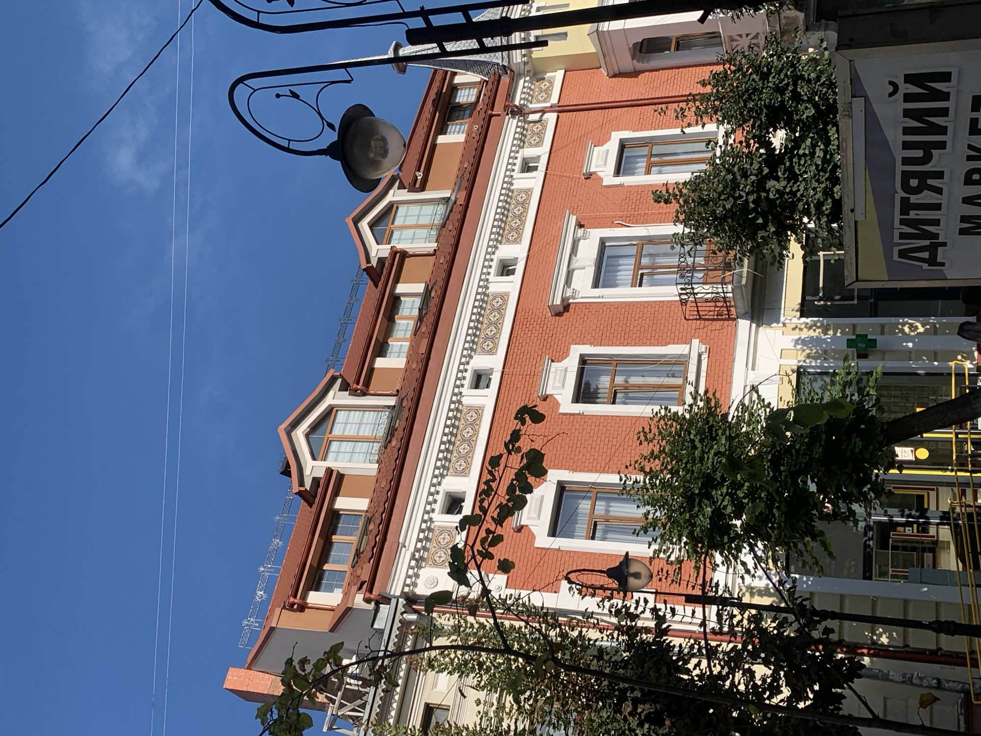 The image depicts a red brick building with a mansard roof, featuring dormer windows. The building is located in Kolomyia, Ukraine. The facade of the building is adorned with decorative white trim and patterned tiles. A black wrought-iron balcony is visible on the second floor. Green trees and streetlights are present in the foreground, partially obscuring the building. A sign with Cyrillic lettering, reading "ДИТЯЧИЙ MARKET," is visible at the bottom of the image. The sky is clear and blue.