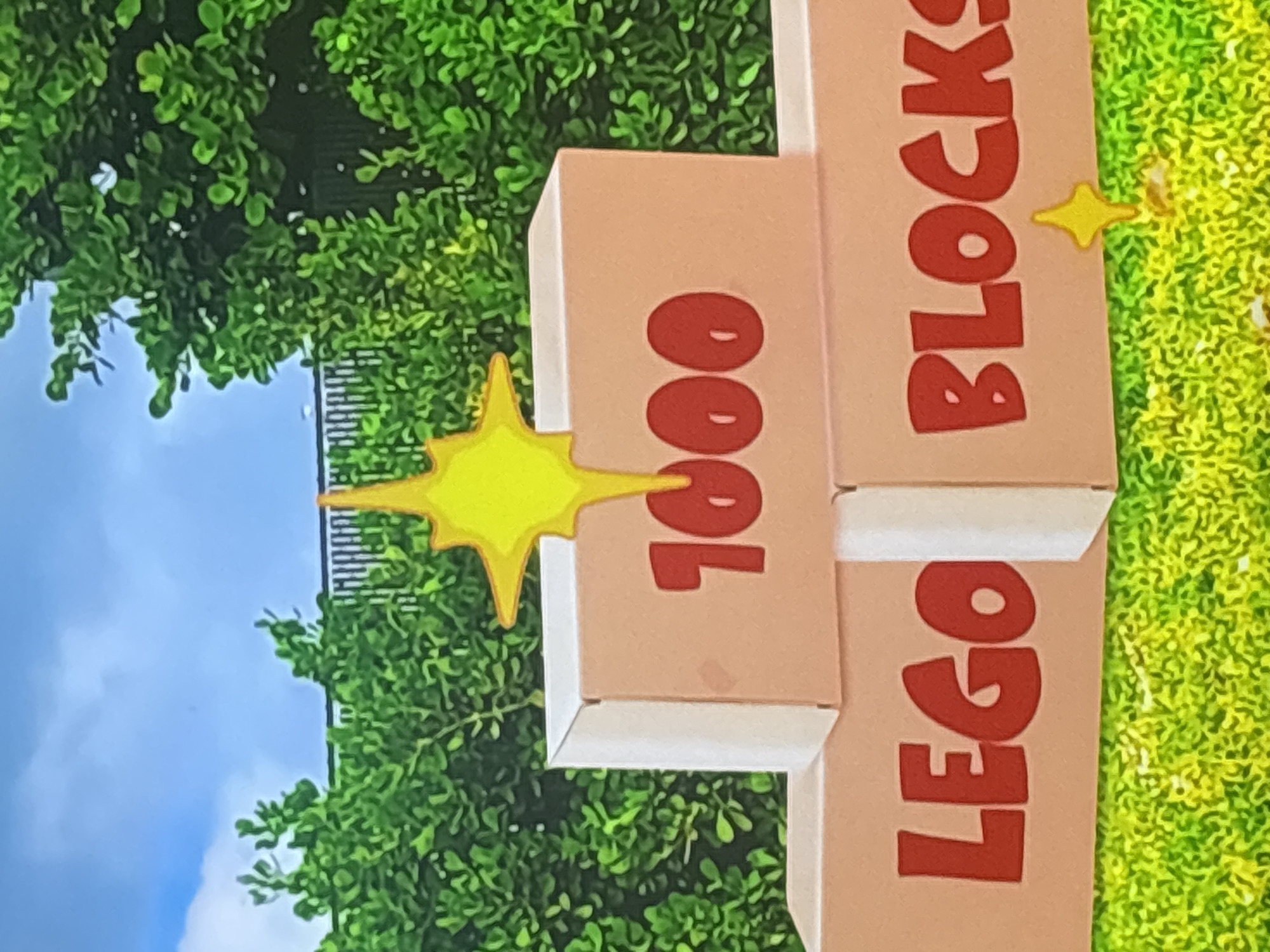 The image shows three stacked cardboard boxes with the text "1000 LEGO BLOCKS" written in red lettering. The boxes are arranged in a pyramidal shape, with one box on top and two boxes below. A yellow starburst decoration is placed on top of the upper box. The boxes are situated outdoors, on a green, grassy surface. In the background, there is a lush green hedge and a clear blue sky. The scene appears to be a decorative display. There are no people or specific landmarks that would indicate a location in Dubai, United Arab Emirates.