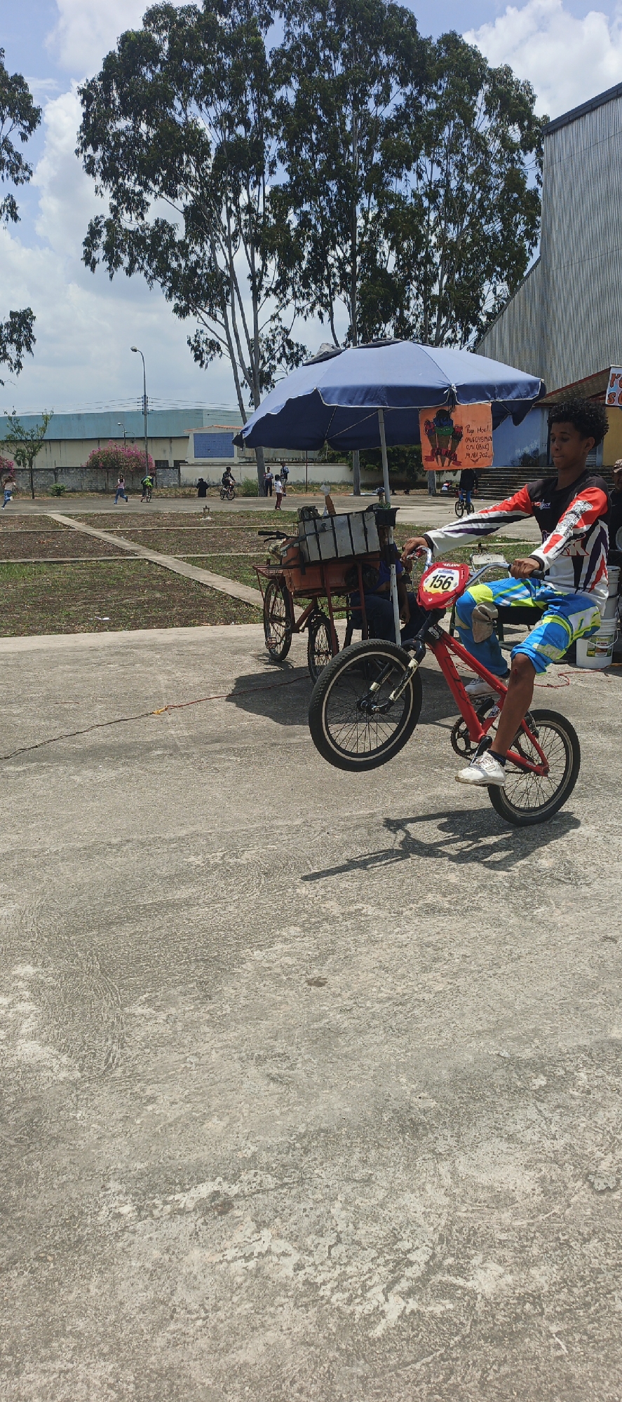 The image captures a young male in Tinaquillo, Venezuela, performing a wheelie on a red bicycle marked with the number "156." He wears a racing jersey and blue and yellow pants with white shoes. In the background, there is a food cart under a blue umbrella, and a group of people is visible in the distance. The setting appears to be a public space, possibly a park or recreational area.
