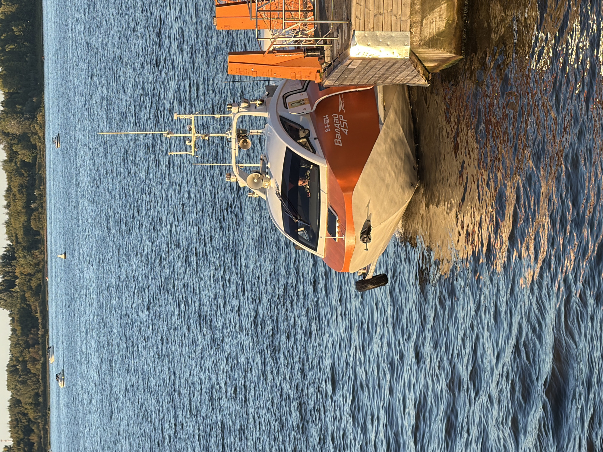 A small, orange and white motorboat, identified by Cyrillic text on its hull as "Валдай 45Р" and a registration number "В-8-6284", is docked next to a wooden pier on a body of water. A person is visible inside the boat's cabin. The water is choppy and reflects the warm light of the sun, suggesting it is either sunrise or sunset. The background features a tree line with a few boats in the distance and a clear sky. The pier has orange safety equipment attached. The scene is likely located on the Volga River in Nizhny Novgorod, Russia, given the type of boat and the general landscape often seen along its banks.