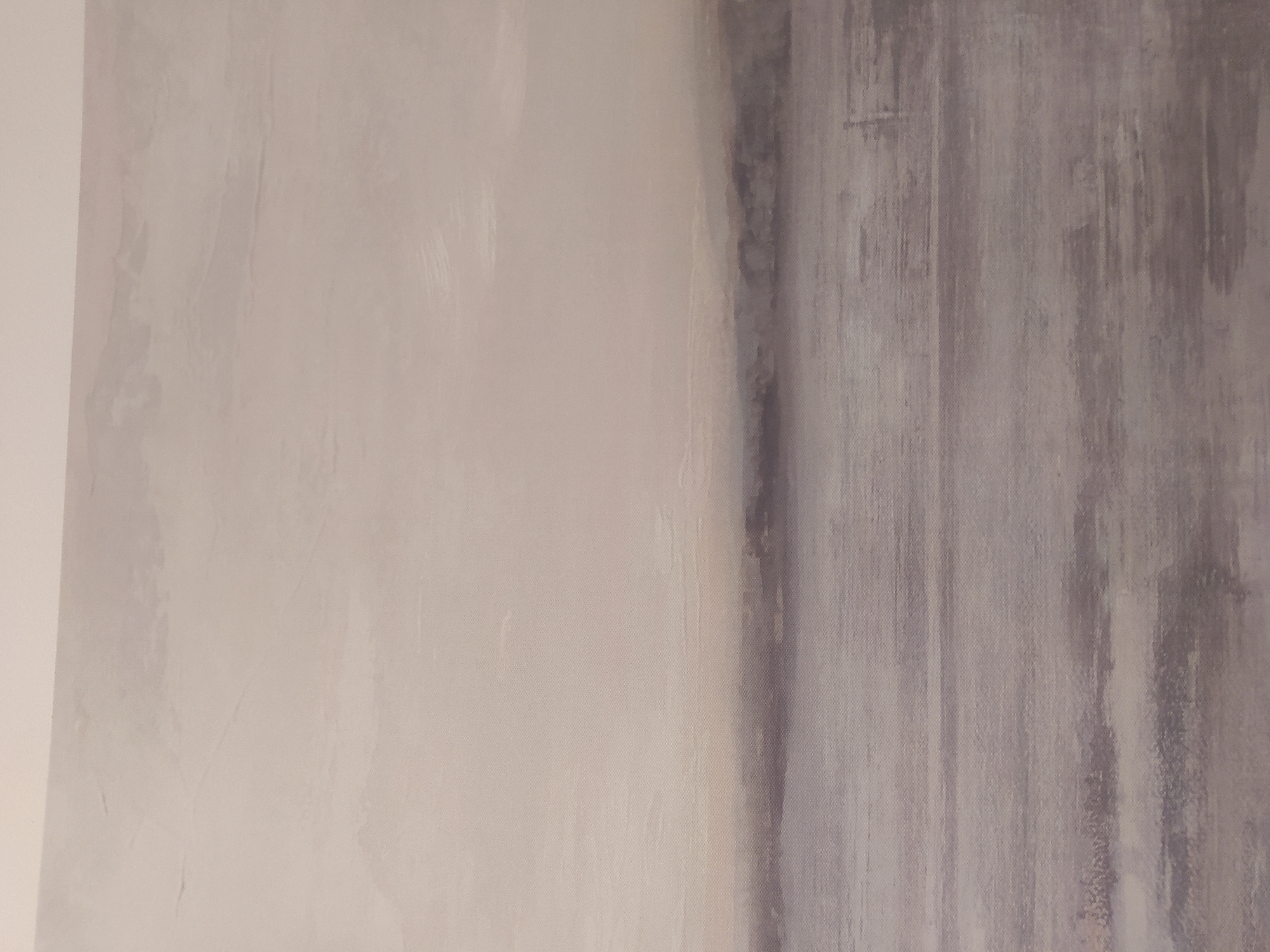 This is a photograph of an abstract painting. The painting features a muted color palette dominated by shades of beige, grey, and white, creating a serene and atmospheric depiction. The top portion of the painting consists of a pale, textured expanse, suggestive of a sky or clouds. Below this, a blurred, horizontal band of darker grey and muted blues represents a distant landscape or horizon line. The bottom two-thirds of the painting is filled with a representation of water, rendered in horizontal strokes of varying shades of grey and hints of lighter tones, creating a sense of depth and movement. The overall style is impressionistic and minimalist, with soft edges and a focus on texture and color.