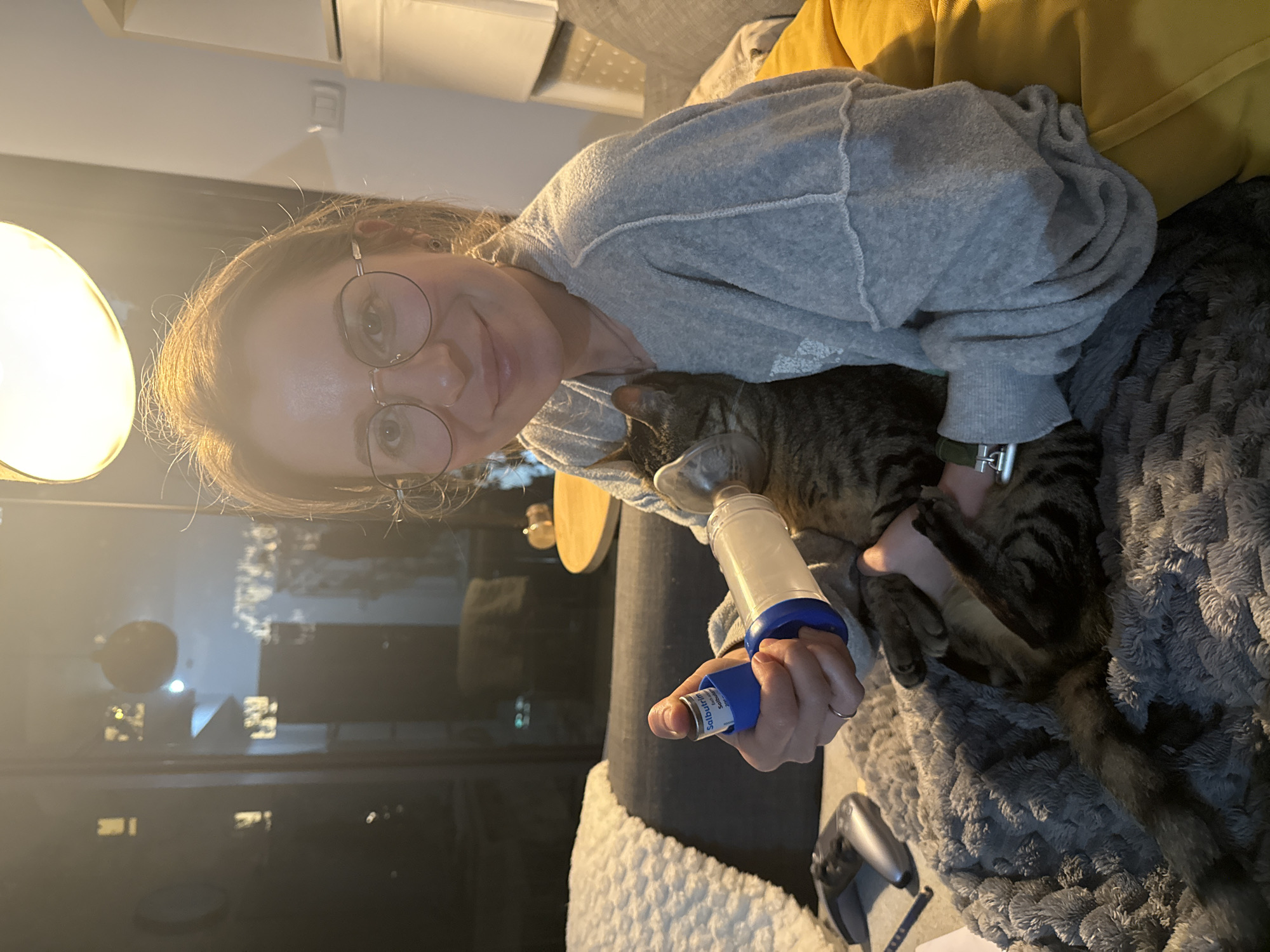 A woman with round glasses and a gray sweatshirt is holding a cat on a gray blanket. She is administering medication to the cat using an inhaler with a blue nozzle and a cylindrical spacer. A yellow pillow is behind her. In the background, there is a window reflecting city lights. The location is Bat Yam, Israel.