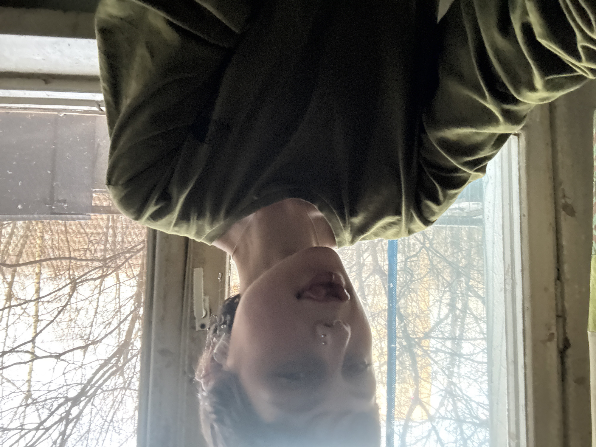 The image shows a person from the chest up, head upside down, wearing a green shirt. The background is a window with trees outside, suggesting an indoor setting. The window appears old with possibly a screen and visible frame. Specific details indicating the location as Moscow, Russia, are not present in the image.