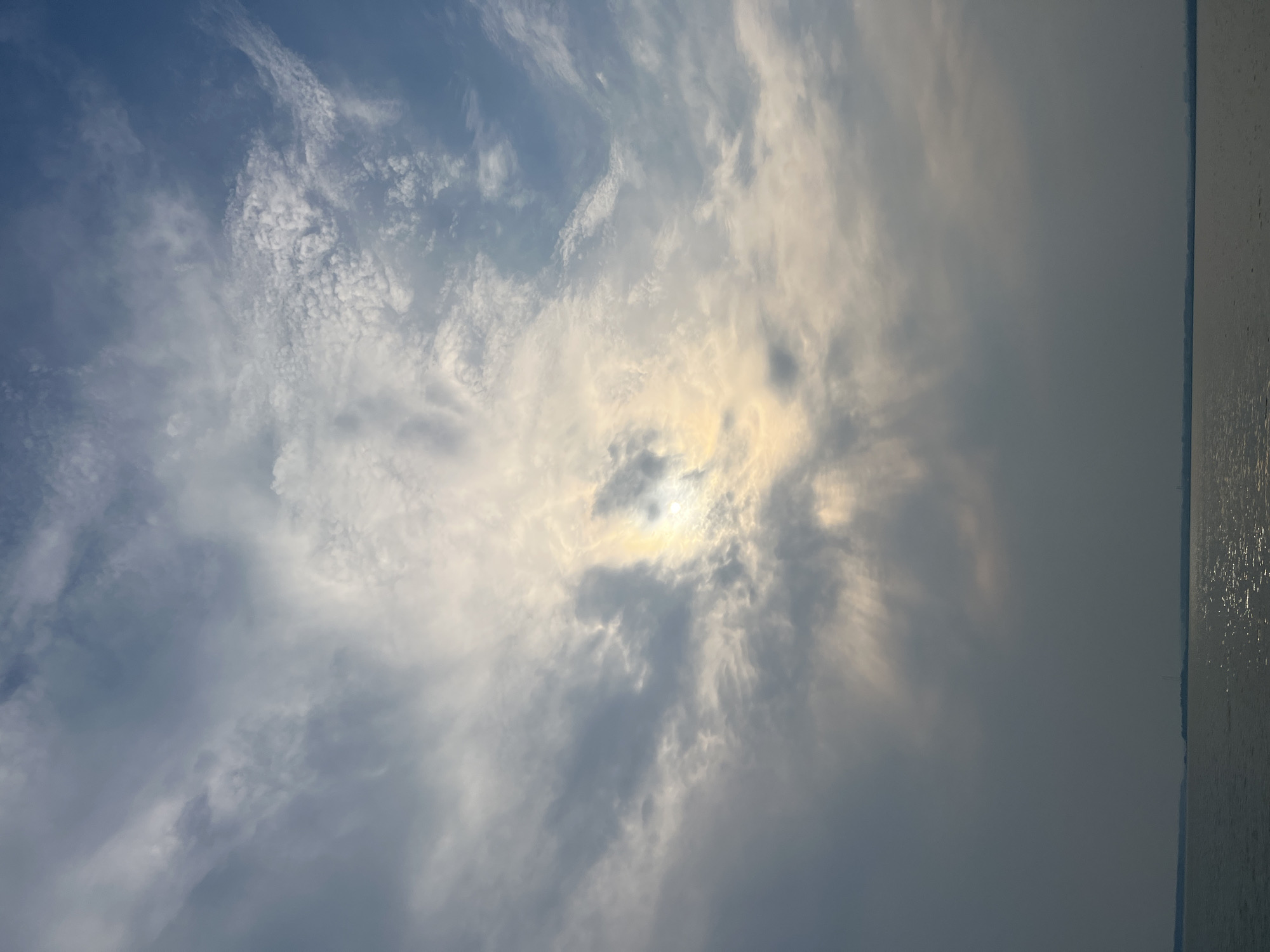 The image shows a body of water, likely a river or lake, with a distant shoreline under a cloudy sky. The sun is partially obscured by clouds, creating a luminous effect with rays of light filtering through. The water reflects the sky and sunlight, creating shimmering patterns on its surface. The sky is a mix of blue and gray clouds, with wispy formations suggesting movement. The shoreline appears flat and featureless, with some faint indications of distant structures or vegetation. The overall impression is one of a natural landscape, possibly a waterfront view in the city of Novopichugovo, Russia.