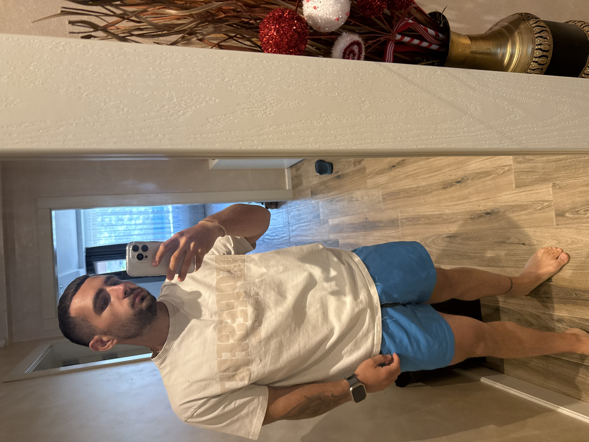 A man is standing and taking a selfie in what appears to be a hallway or a room with wood-paneled flooring. He is wearing a light-colored t-shirt with an embossed design on the front and blue swim shorts. He has a watch on his left wrist and is barefoot. In the background, a decorative vase with red and white ornaments is visible. The lighting suggests an indoor environment. The image does not contain any specific visual cues that would allow for determination of the location within Gabrovo, Bulgaria.