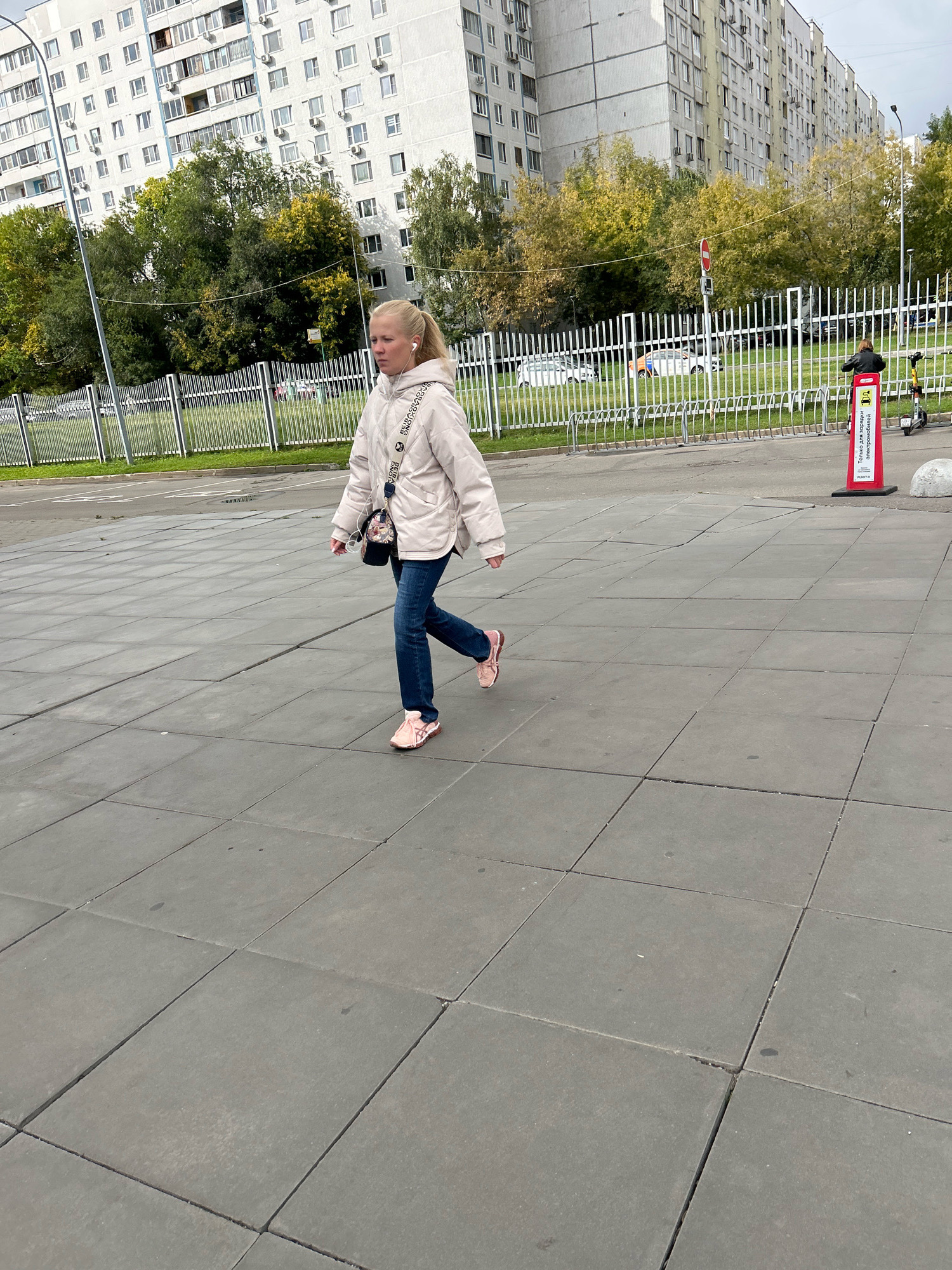 A young woman with blonde hair pulled back in a ponytail walks on a paved area in front of a tall residential building. She is wearing a light-colored hooded jacket, dark blue jeans, and pink sneakers. She has a small purse with a floral pattern slung across her body and is wearing white earbuds. In the background, there are trees with yellowing leaves and a white metal fence. Beyond the fence, several cars are visible. A red sign with white text is positioned to the right of the woman, next to a scooter rental station. The residential building in the background is typical of Soviet-era architecture, with numerous windows and balconies. The scene appears to be a public space in a residential district of Moscow, Russia, likely on an overcast autumn day.