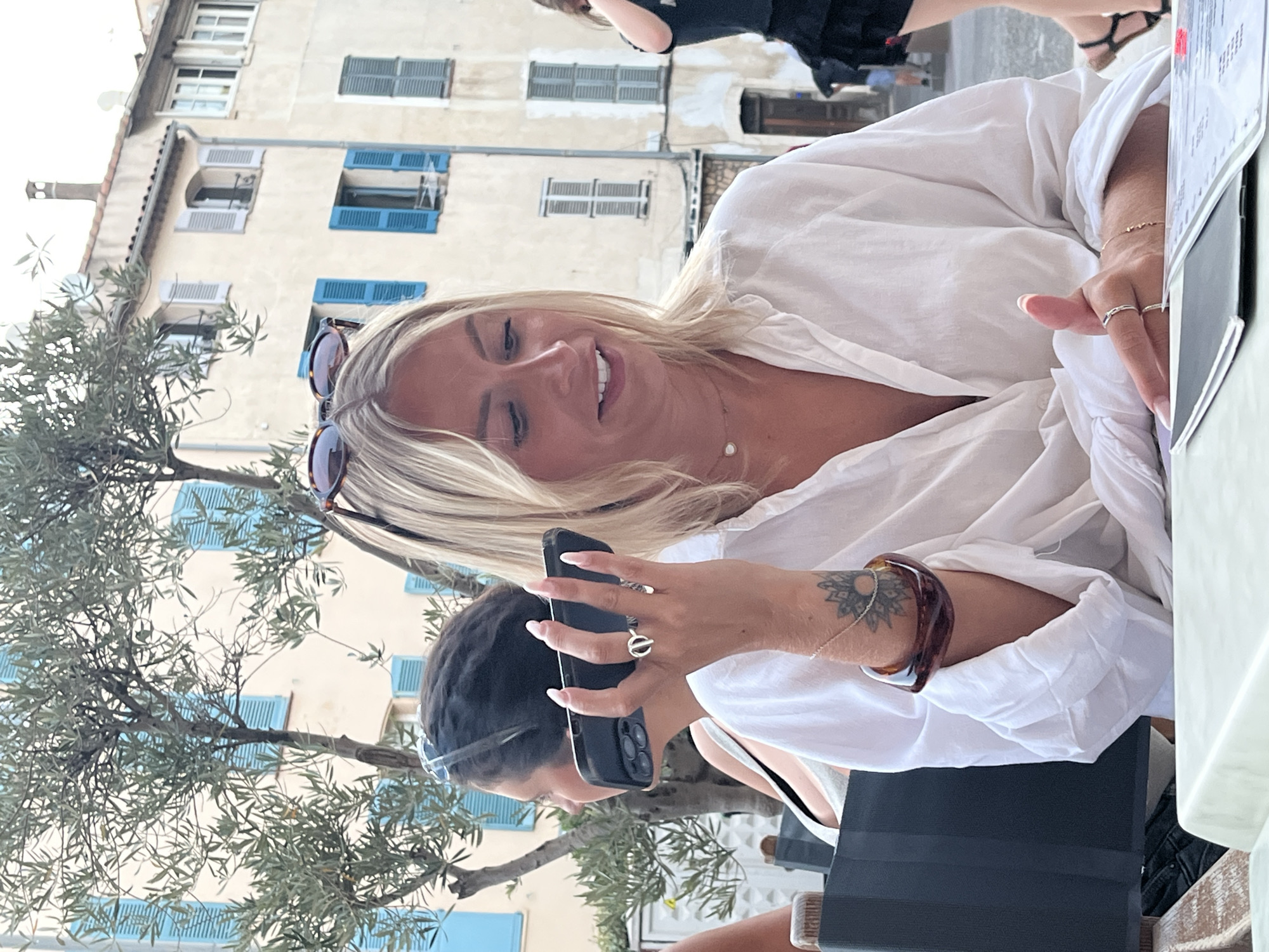 The image shows a close-up of a blonde woman sitting at an outdoor cafe. She is wearing a white linen shirt and looking at her phone. She has sunglasses on her head and is wearing rings on her fingers and bracelets on her arm. Behind her, the facade of a building with blue shutters is visible, as is a tree with green leaves. The scene appears to be set in Antibes, France, judging by the architecture and the presence of an olive tree. The lighting suggests it is daytime, possibly late afternoon given the warm glow. The woman's expression is relaxed and engaged with her phone, indicating a casual moment.