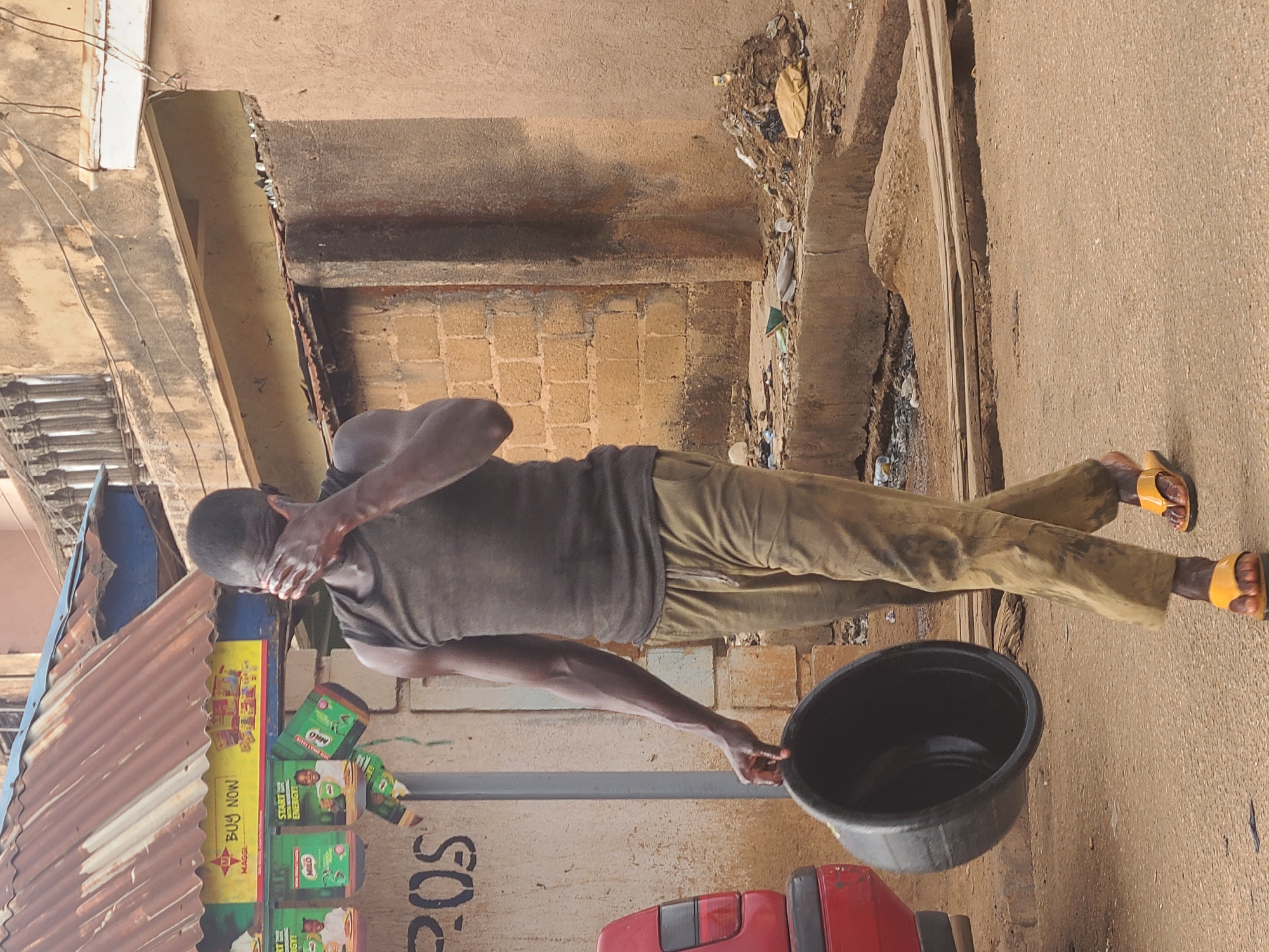 A man stands on a street in Jos, Nigeria. He is wearing a dark tank top and light brown pants with yellow sandals. He is holding a black bucket in his left hand and covering his face with his right hand. Behind him is a building with a corrugated metal awning and advertisements for Milo. The building is painted with the letters "POS". The street is made of dirt. A red vehicle is partially visible in the lower right corner. The overall environment appears to be urban and somewhat impoverished.