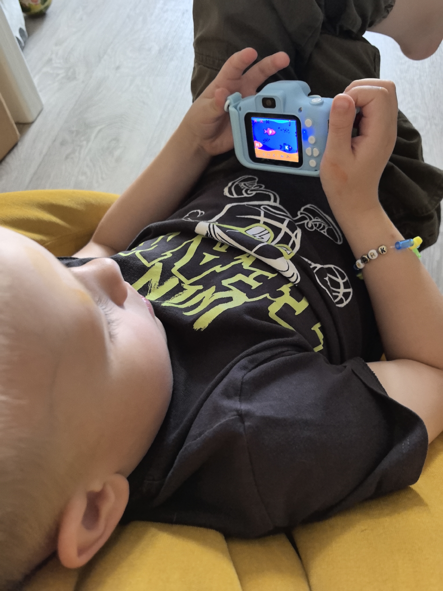 An indoor, eye-level shot captures a young child lying down and engrossed in a light blue digital toy camera or gaming device. The child is positioned on a soft, yellow surface, likely a cushion or sofa, with their head resting to the left and looking towards the right. Their short, light hair and a portion of their face, including the forehead and eye area, are visible, suggesting a moment of focused attention.
The child wears a dark t-shirt adorned with a vibrant lime green and white graphic design, which appears to be a stylized pattern or abstract text. Their lower body is covered by dark green or olive-colored pants, visible from the mid-torso downwards. Both hands hold the blue device, which features a small screen displaying an animated underwater scene with two red fish and the word "SHRIMP" at the bottom. Several white control buttons are visible on the right side of the device, implying interactive play.
A notable detail is a multi-colored beaded bracelet on the child's right wrist. White letter beads clearly spell out "MARK," interspersed with other colorful beads, and a small blue and yellow charm resembling a toy water gun. In the blurred background to the top left, a segment of light-colored wooden laminate flooring is visible, alongside an out-of-focus object that could be furniture or another toy.
The child appears to be actively engaged in playing with the device, indicating a moment of entertainment. The lighting suggests it is daytime. The scene is set indoors, likely within a home environment in Perm, Russia.