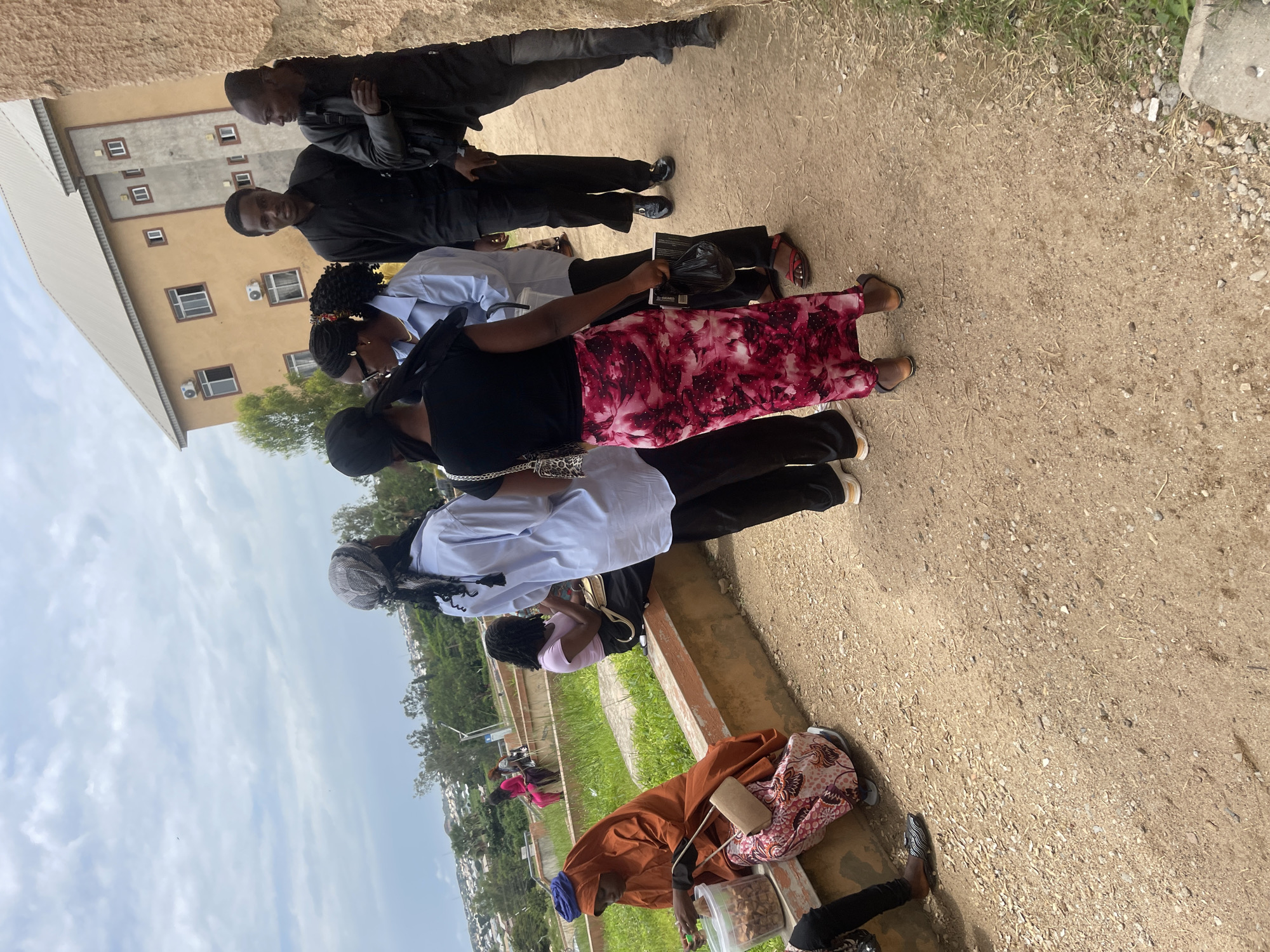 The image shows a group of people standing and sitting outside a building in what appears to be a dusty, outdoor urban environment. The building in the background is a multi-story structure with a light brown facade and beige trim, featuring several windows and what look like air conditioning units. The ground is covered in dirt and small pebbles.
In the foreground, several individuals are visible. On the far left, a woman wearing an orange hijab and a patterned skirt is sitting on a low wall, holding a clear container with some items inside. Next to her, a person in black pants and a black and white striped shoe is partially visible.
A group of people are gathered in the center, facing each other. One person wears a light blue shirt and black pants, another a black shirt and black pants, and a third a black shirt with a pink and red floral skirt. The woman in the floral skirt is wearing sandals. Behind them, a person in a black jacket and black pants is standing.
Further back, other individuals are seated on the same low wall. Two women are visible, one in a bright pink outfit and another in a light pink shirt.
The environment suggests an area in Jos, Nigeria, with the architecture of the building and the style of dress being consistent with the region. The overall impression is a casual gathering or waiting area outdoors.