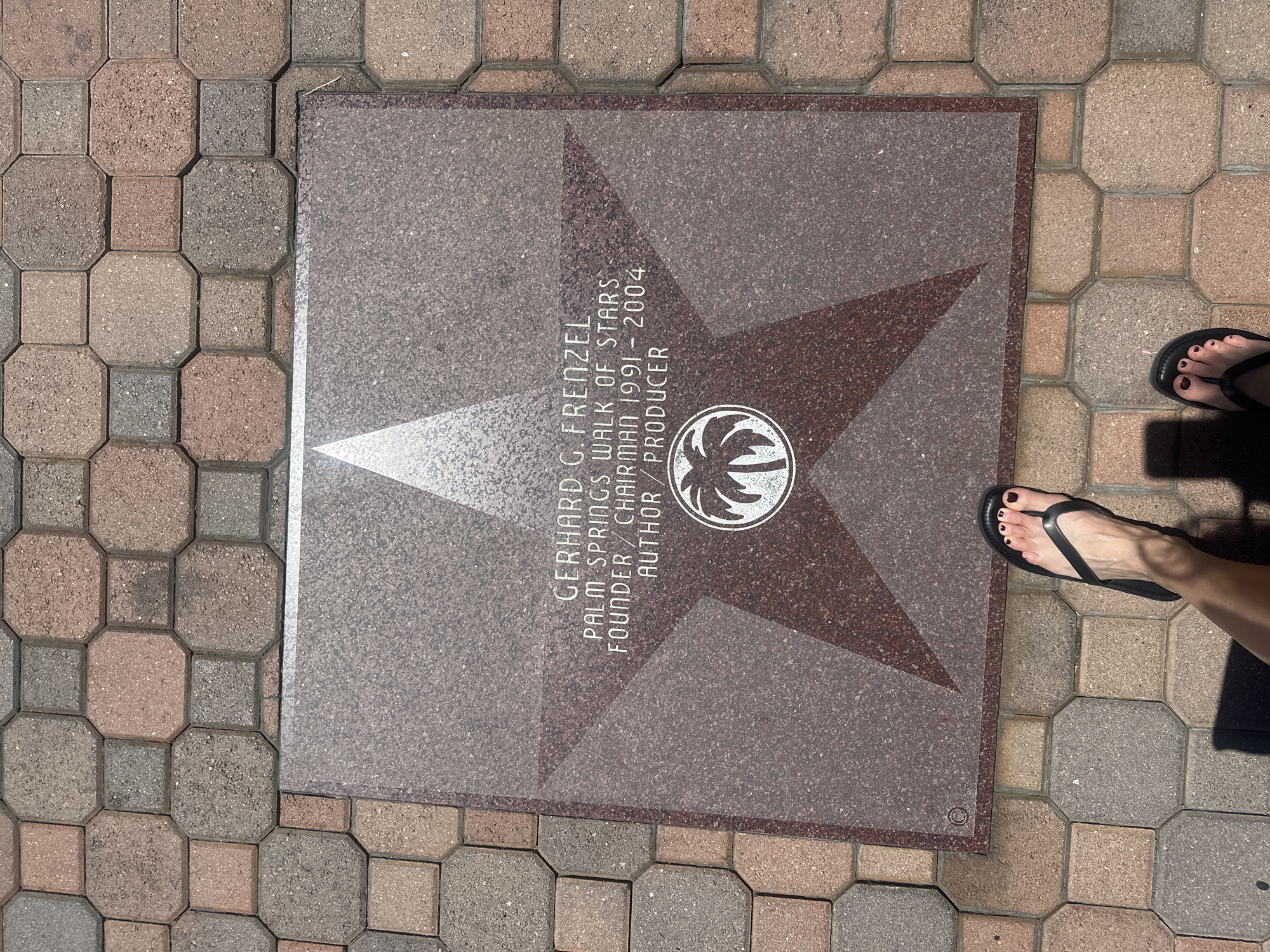 The image depicts a stone plaque set into a brick-paved walkway. The plaque is square-shaped and features a five-pointed star design. Within the star is a palm tree emblem. Inscribed on the plaque are the words "GERHARD G. FRENZEL, PALM SPRINGS WALK OF STARS, FOUNDER/CHAIRMAN 1991-2004, AUTHOR/PRODUCER". A pair of feet wearing black flip-flops are positioned next to the plaque. The setting is outdoors.