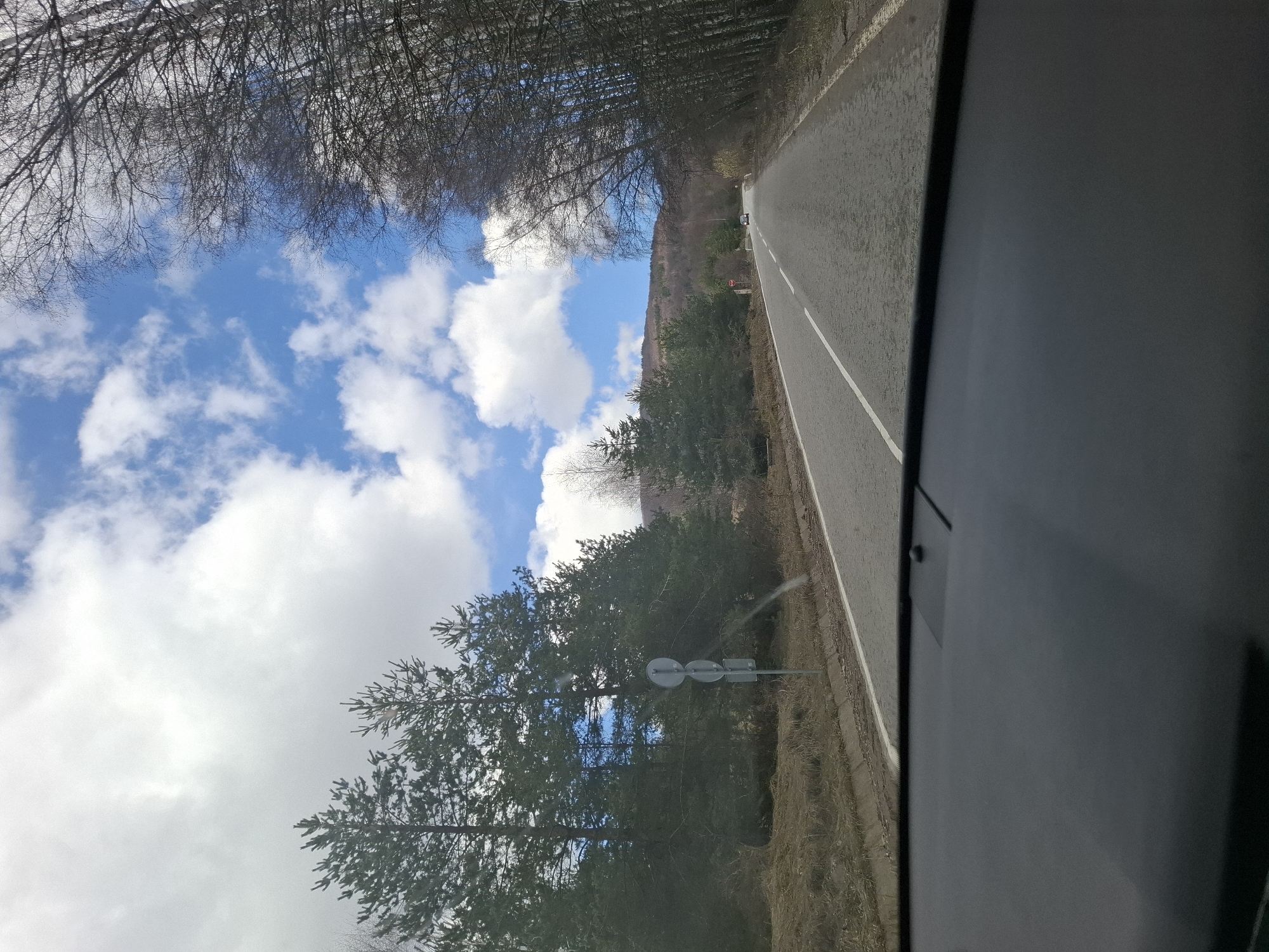 The image shows a view from inside a vehicle, likely a car, traveling on a road in Gintsi, Bulgaria. The road has a solid white line dividing the lanes. To the left of the road is a mix of trees and foliage. A road sign is visible alongside the road. In the distance, a vehicle is moving along the road. Above the treeline, the sky is visible with a mix of blue sky and white clouds. The overall scene suggests a drive through a somewhat rural or forested area.