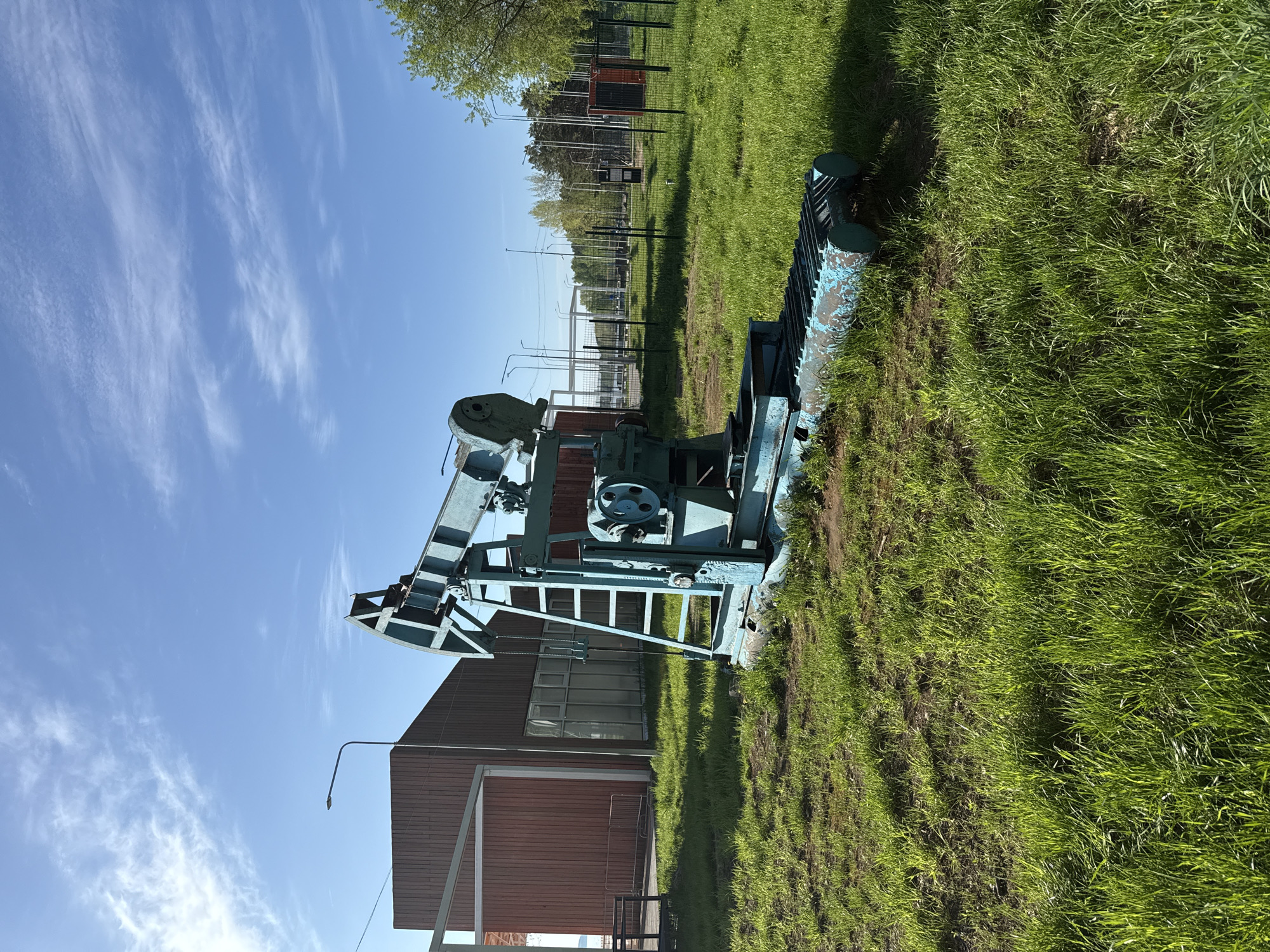 The image depicts an oil pumpjack on a grassy field in Chaikovsky, Russia. The pumpjack is painted blue and positioned next to a red wooden building. The sky is blue with wispy clouds. A fence line and trees are visible in the background.