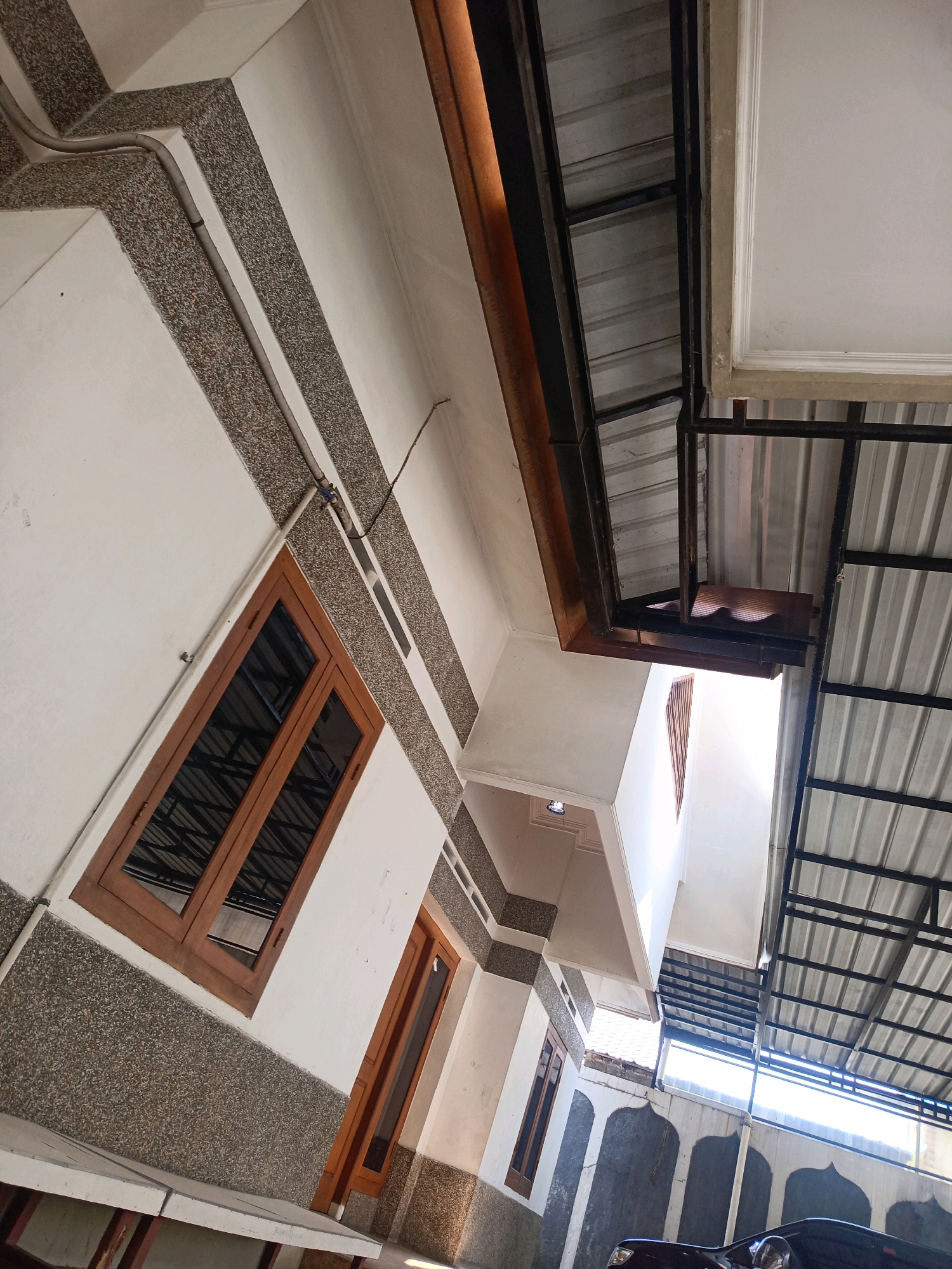 The image shows an angled view of an exterior building facade and a covered outdoor area, likely a carport or patio. The building features white walls with horizontal bands of speckled, textured material. Two wooden-framed windows are visible on the building's side, one larger and one smaller. A wooden door is also present. Above and to the right, a metal roof structure with corrugated metal sheeting is supported by dark metal beams, casting shadows. Sunlight streams through an opening in this structure, illuminating the area below. In the lower right corner, a dark-colored car is partially visible. The architectural style suggests a residential or commercial building. The overall scene is devoid of people and activity. This scene is located in Magetan, Indonesia.
