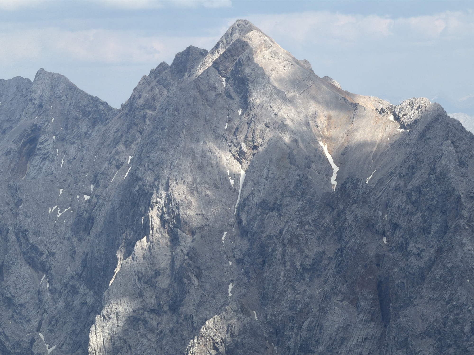 This media file is a photograph of a mountain range, likely taken in the vicinity of Garmisch-Partenkirchen, Germany. The primary subject is the rugged, rocky terrain of several peaks, with one prominent, sharp peak dominating the center of the frame. The mountains are composed of gray, weathered rock, with visible striations and crevices. Patches of snow are scattered across the slopes and in some of the shaded gullies, indicating a cool climate or high altitude.
The lighting suggests it is daytime, with the sun casting distinct shadows and illuminating parts of the mountainside. The light creates a dramatic contrast between the sunlit areas, which appear lighter and almost golden in hue, and the shadowed areas, which are a deep, cool gray. The sky above is partly cloudy, with soft white clouds against a pale blue background, indicating fair weather with some atmospheric haze.
There are no people or man-made objects clearly visible in the image. The scene is one of natural, untamed wilderness, emphasizing the raw beauty and imposing scale of the mountains. The texture of the rock is highly detailed, allowing for an appreciation of the geological formations. The overall impression is one of majesty and solitude. No visible text is present in the image.
