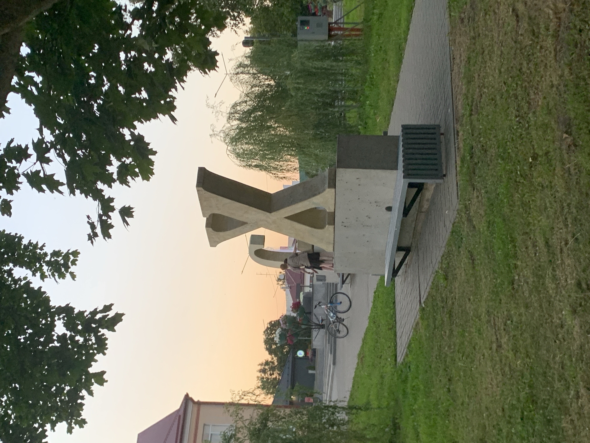 This is a medium shot, outdoor photograph taken at ground level. The scene depicts a large, gold-colored letter 'X' sculpture, mounted on a concrete base. To the left of the sculpture, a bench with a slatted metal front is visible. Behind the sculpture, two people are standing, one of whom is leaning on the sculpture. A bicycle is parked on a paved pathway to the left of the sculpture and bench. The background includes a building with a red roof to the far left, trees, and a weeping willow tree. The sky is a pale orange, suggesting it is either sunrise or sunset. The photograph was taken in the city of Kapyĺ, Belarus.