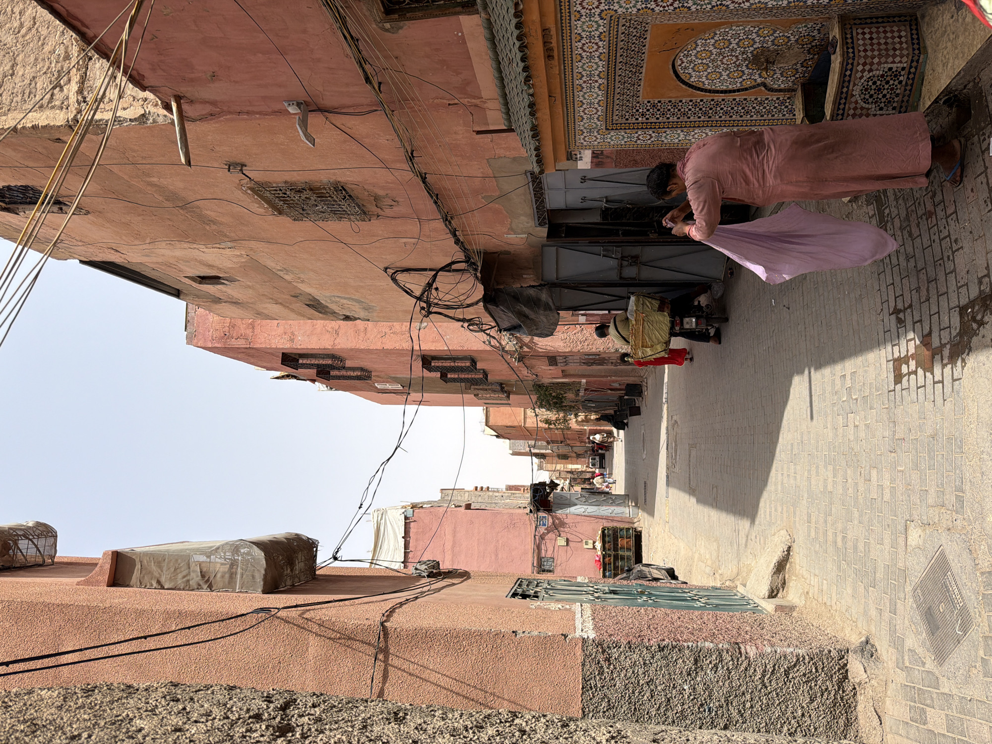 The image depicts a narrow, sun-drenched alleyway in the Marrakech Medina, Morocco. The buildings lining the alley are made of reddish-pink stucco, with some showing signs of age and weathering. The architecture is typical of the old city, featuring small, barred windows and ornate tilework on a fountain to the right of the frame.
In the foreground, a person wearing a long, light pink garment and sandals is holding up a lavender-colored fabric. To their left, a motorcycle with a large basket is parked, and another person is partially visible, appearing to push it. Further down the alley, other people are walking, some dressed in traditional Moroccan attire. The ground is paved with cobblestones, and shadows are cast by the buildings, indicating it is midday. Overhead, a tangle of electrical wires is visible against the pale blue sky. The scene conveys a sense of everyday life in the bustling, historic heart of Marrakech.