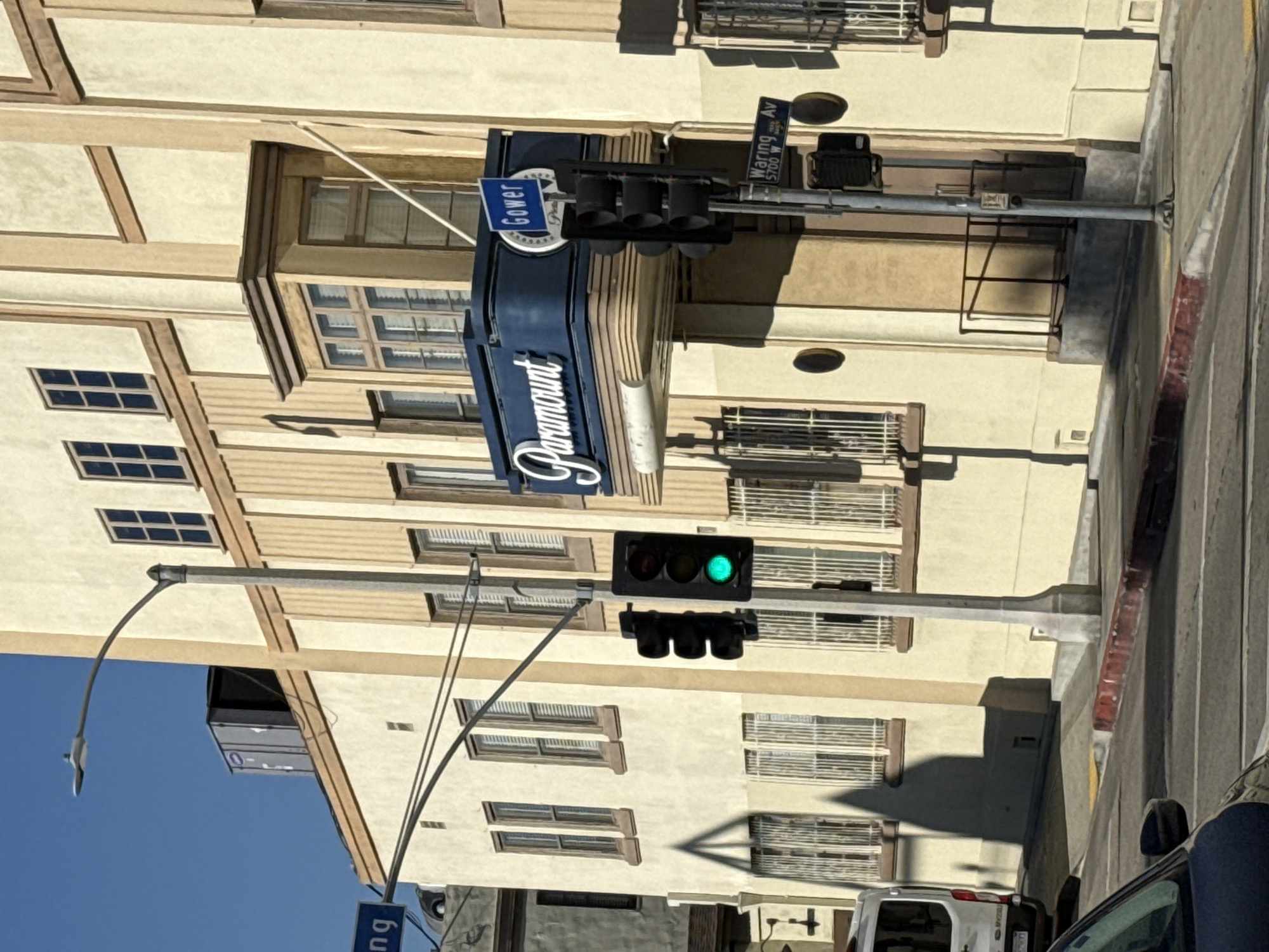 The image depicts a street scene in Los Angeles, United States, dominated by the Paramount Pictures studio building. The building is beige, with dark-framed windows. A large, curved blue sign displaying the "Paramount" name hangs on the building's corner. Above the sign, there's a street sign indicating "Gower" and another sign reads "Waring Av 5700 W." A traffic light is visible, displaying a green light. The sky is clear and blue. A car is partially visible in the bottom right corner, and a white van is parked on the street in the lower portion of the image.