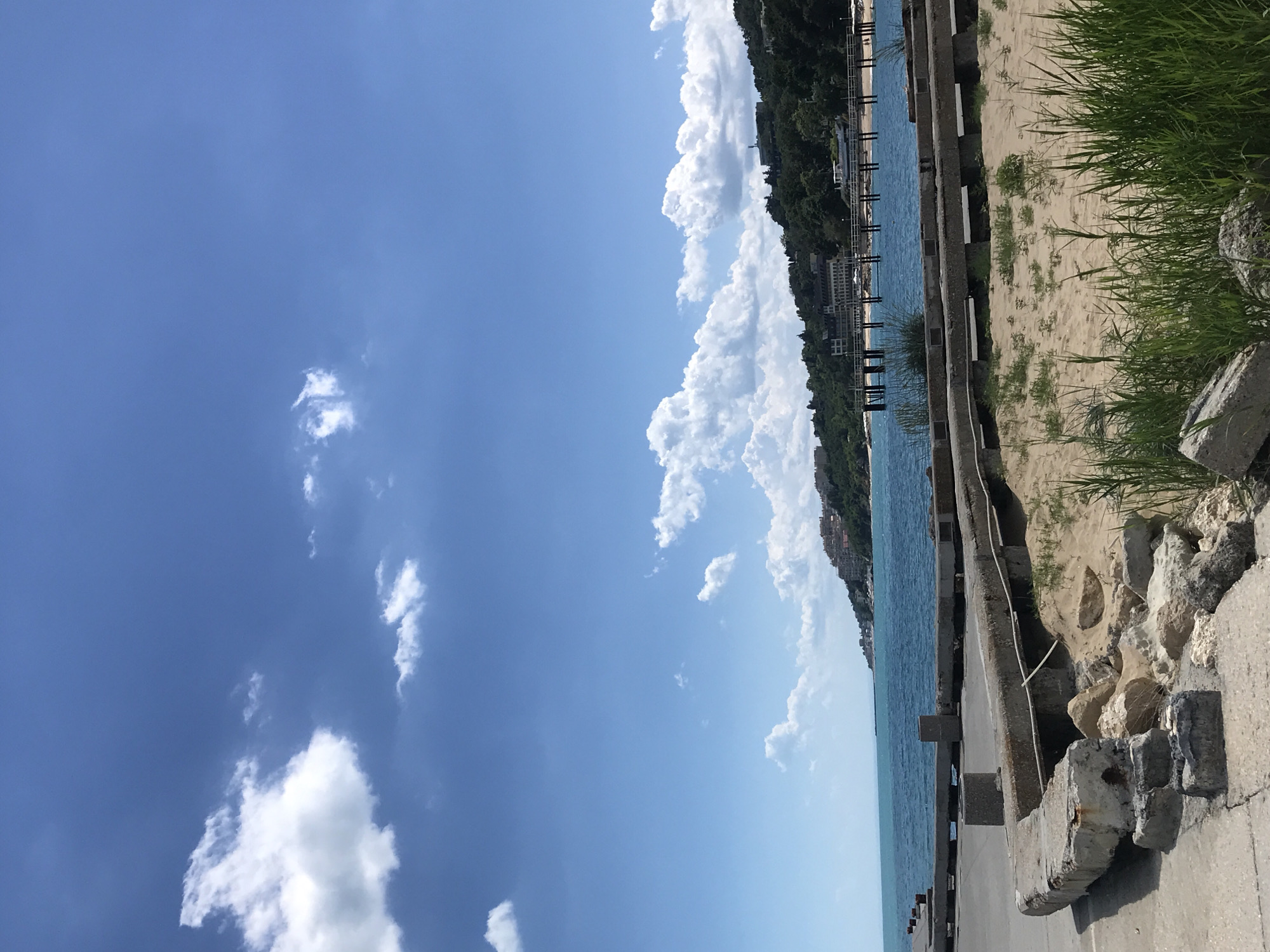 The image shows a view of the Black Sea coast in Varna, Bulgaria, on a sunny day with a bright blue sky and scattered white clouds. In the foreground, there is a cracked concrete walkway and sandy area with some rocks and green grass. Beyond this, the turquoise sea stretches out. A wooden pier extends into the water, with a beach visible on the far side, backed by a densely wooded hill. Buildings, some appearing to be hotels or resorts, are visible nestled amongst the trees on the hillside. The overall scene depicts a coastal resort area.