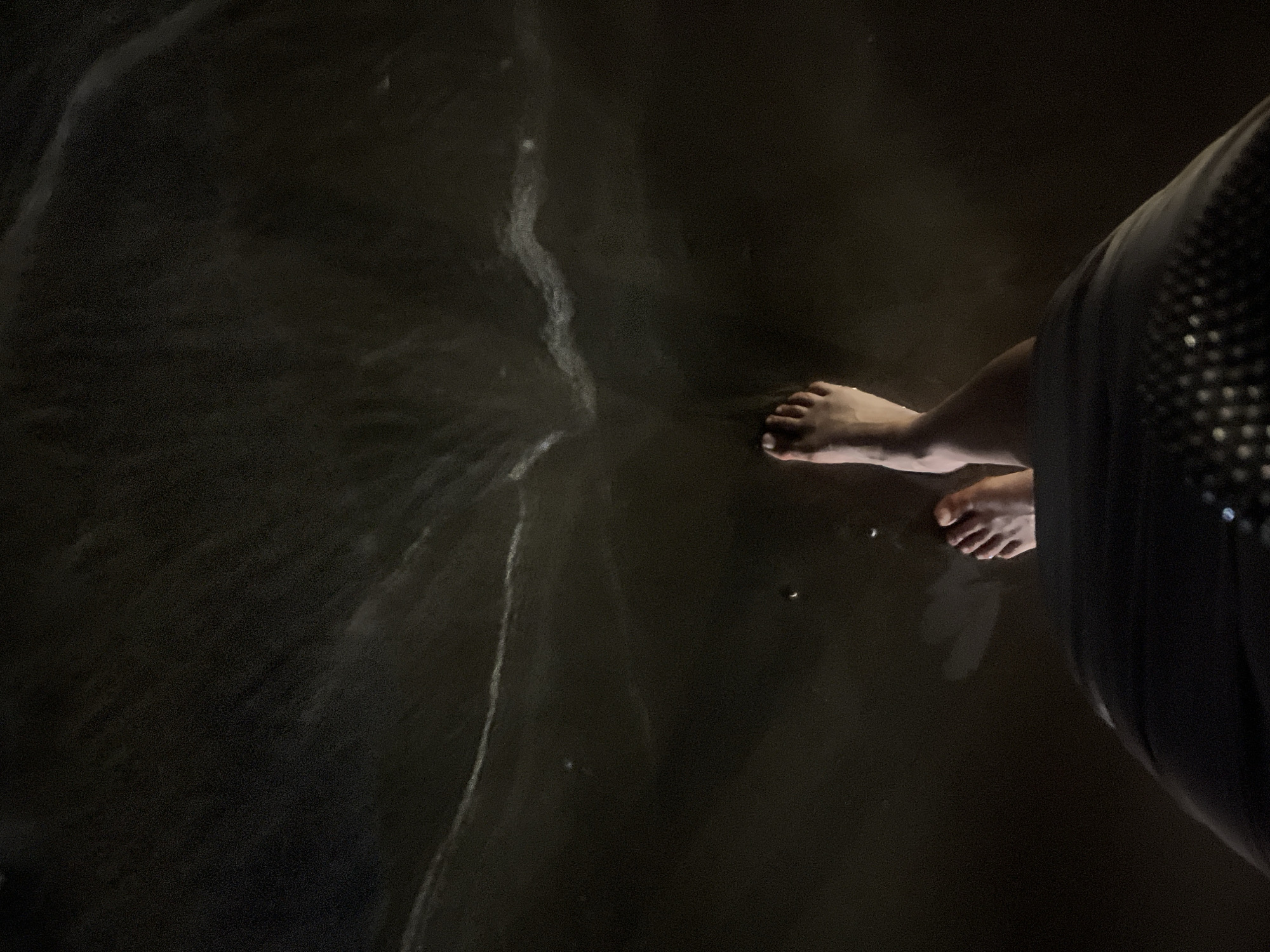 The image presents a high-angle view looking down at a person's bare feet in dark water, likely at night. The feet are positioned towards the right side of the frame, with the left foot slightly ahead of the right. The skin tone of the feet appears light, contrasting with the dark surroundings. A portion of the person's lower leg and dark clothing is visible on the far right edge of the frame. The clothing, possibly a skirt or dress, exhibits subtle shimmering or reflective details.
The scene depicts water that is almost black, suggesting either very low light conditions or dark sand beneath shallow water. Faint, ethereal white lines and streaks are visible on the water's surface, indicating reflections of light or the movement of water/foam. The overall lighting is very dim, strongly implying the photograph was taken at night. Given the location context of Đà Nẵng, Vietnam, this scene most likely takes place on a beach or shoreline. There are no other discernible objects or details in the background due to the darkness. No text is visible in the media file.