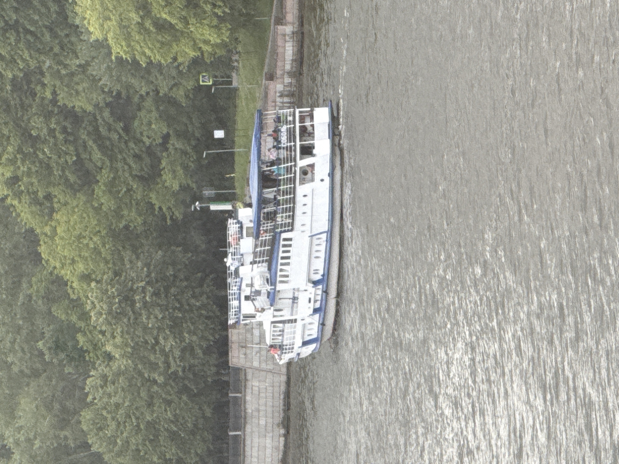 A white double-decker tour boat is moving on a body of water. The boat has a blue canopy covering the upper deck and white railings. There are people on the upper deck. A name that appears to be "СОБОЛ" is visible on the side of the boat in dark lettering. The boat is near a stone embankment with trees in the background. In the distance, there are streetlights and a yellow sign with a pedestrian crossing symbol. This scene is consistent with a tour boat on the Moskva River in Moscow, Russia. The water appears choppy.