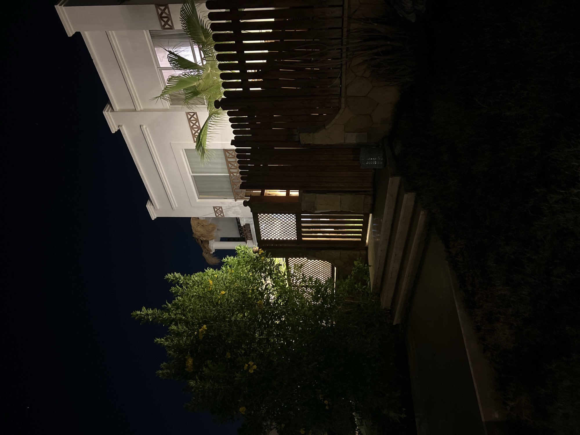 The image depicts a nighttime exterior view of a modern villa in Sharm El Sheikh, Egypt. A lush green tree with yellow flowers is prominent on the left side of the frame, illuminated by an unseen light source. Adjacent to the tree is a wooden gate with a decorative lattice design, leading to a pathway with stone steps. Behind the gate and tree, a section of the villa's facade is visible, characterized by white walls and large windows. A wooden fence with vertical planks borders the property, with a palm tree extending above it. The scene is set against a dark night sky, with a single star visible. The overall impression is one of a well-maintained private residence at night.