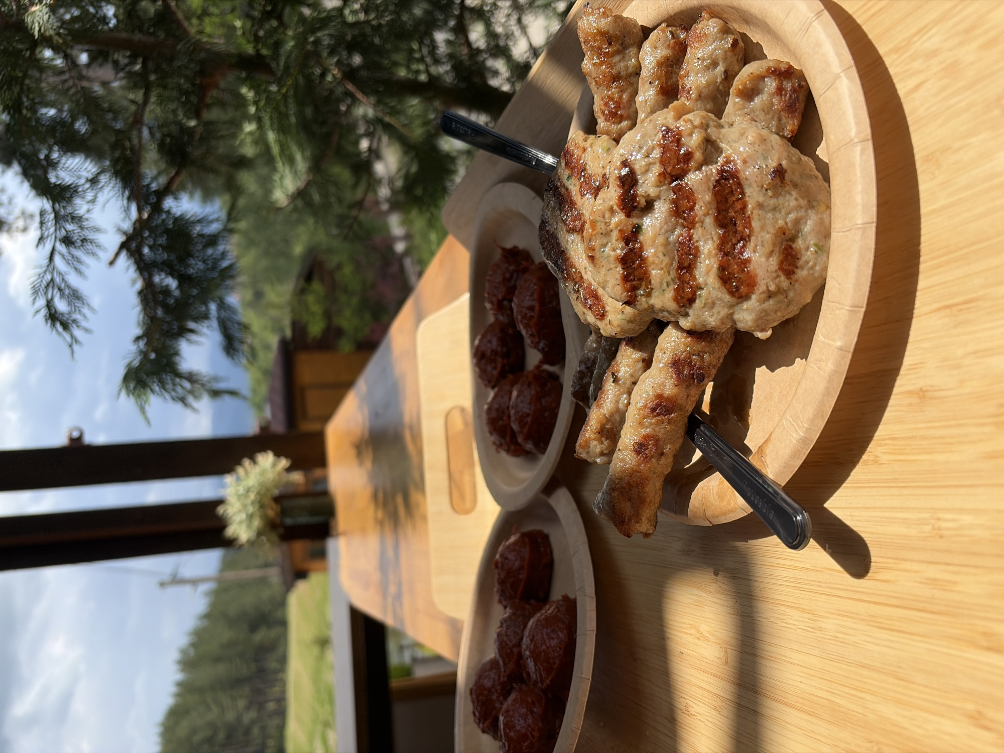 The scene depicts an outdoor dining arrangement on a wooden table, set in Alino, Bulgaria. In the foreground, a light-colored, possibly bamboo, slatted table is illuminated by direct sunlight, casting distinct shadows from the left. A primary disposable plate in the lower right contains grilled meat: two flattened, oval-shaped patties with prominent char marks, consistent with *kyufte*, surrounded by multiple smaller, elongated, grilled meat cylinders resembling *kebapche*. A black-handled metallic fork is inserted into one of the smaller meat items. Two additional, smaller disposable plates are positioned further back on the table to the left, each holding several individual portions of a dark red, viscous substance, possibly *lyutenitsa* or a similar relish.
Beyond the dining table, a second wooden surface is visible, supporting a small, dark vase containing a cluster of white flowers or foliage, appearing slightly out of focus. The background reveals a natural landscape under a partly cloudy sky. To the left, a dense evergreen forest covers a series of hills, bordering a vibrant green field or clearing. A large, out-of-focus coniferous tree occupies the right side of the background, partially obscuring the view. Distant, hazy mountain ranges are discernible on the horizon. A utility pole with associated power lines is faintly visible in the far left of the background, near the edge of the forest.