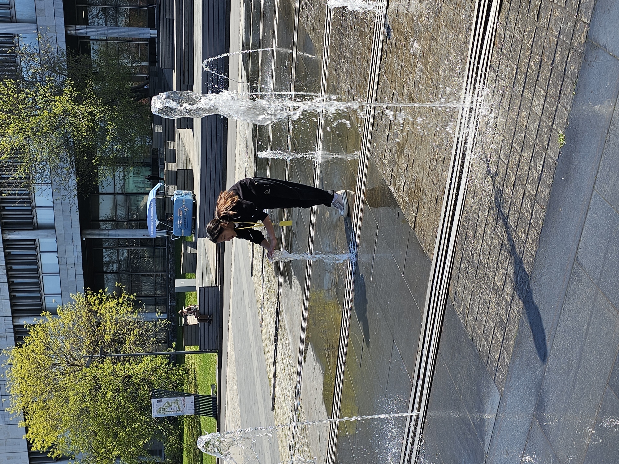 A person wearing a black tracksuit and white sneakers is bending over a water fountain in Moscow, Russia. The fountain is a series of vertical jets of water shooting up from a paved surface. The person has their hands in the water. In the background, there is a modern building, trees, benches, and a blue maintenance vehicle. The scene is outdoors and the lighting indicates it is daytime. The ground is a mix of smooth and textured paving stones. A few people are on benches in the background.