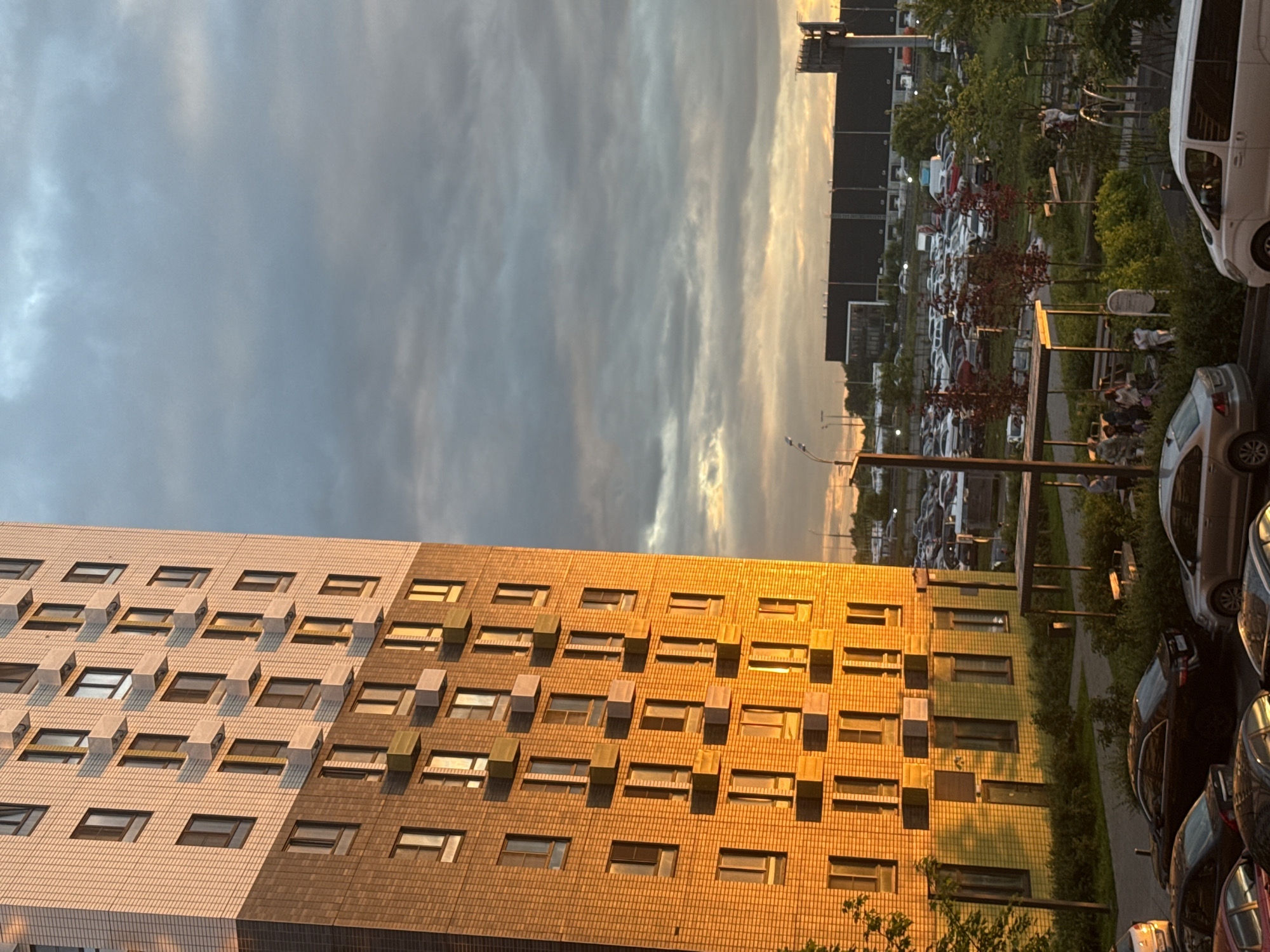 This media file shows an outdoor scene at dusk. The sky is filled with dramatic clouds, transitioning from a muted grey to warmer hues of orange and yellow as the sun sets.
Dominating the foreground is the facade of a modern apartment building, its windows reflecting the fading light. The building's exterior appears to be clad in a mix of light and dark textured tiles. Many of the windows are small and rectangular, some with visible interior structures or reflections. The building stretches across the frame, suggesting a residential complex.
Behind the apartment building, a large parking lot is visible, filled with numerous cars. The cars are parked in rows, and their variety in shape and color adds to the sense of a busy urban or suburban environment. Some trees and greenery are interspersed within the parking area and along the edge of the scene, with some deciduous trees showing red foliage, possibly indicating autumn.
To the right of the parking lot, a white van is parked, its side facing the viewer. Further down, several other cars are parked, including a dark-colored sedan and what appear to be SUVs and hatchbacks. The overall impression is one of a residential area with ample parking.
The lighting suggests it is either sunrise or sunset. Given the warm tones in the sky and on the building, sunset is more likely. The presence of cars and buildings points towards a developed area. No people are clearly identifiable as engaging in specific activities, though there might be figures near the parked cars or within the buildings that are not distinct enough to describe. The scene conveys a sense of the end of the day in a populated, modern setting. No visible text is present in the image.