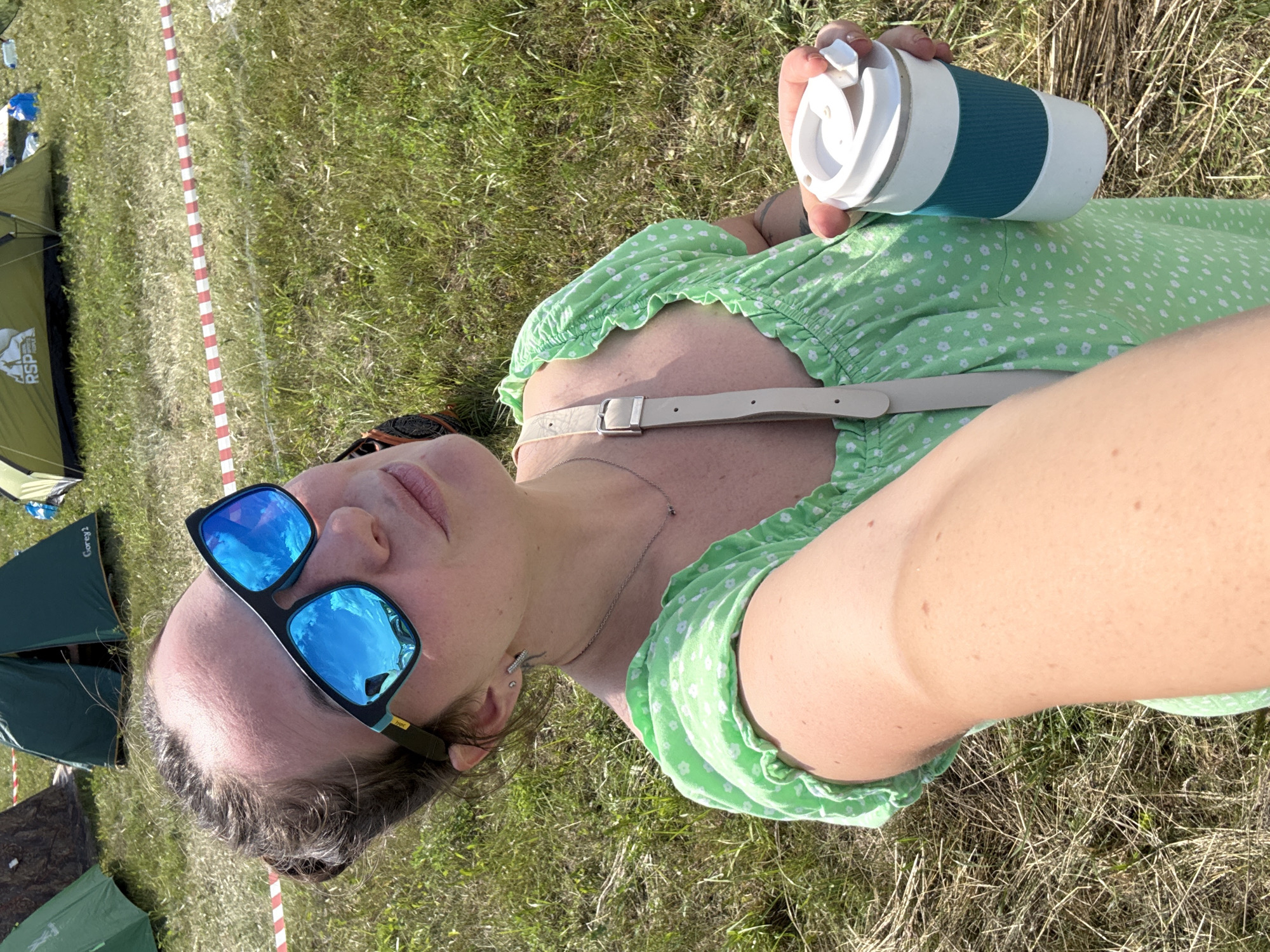 A woman, with her hair pulled up, is taking a selfie in a grassy outdoor area with tents in the background. She is wearing large, blue-tinted sunglasses, a light green dress with a white floral pattern, and a silver necklace. She is holding a white and teal travel mug in her right hand. The environment appears to be a park or campsite, with dry grass and a red and white striped caution tape visible in the background. The scene is outdoors and appears to be during the daytime, with sunlight casting shadows. The location is identified as Lagunovo, Russia.