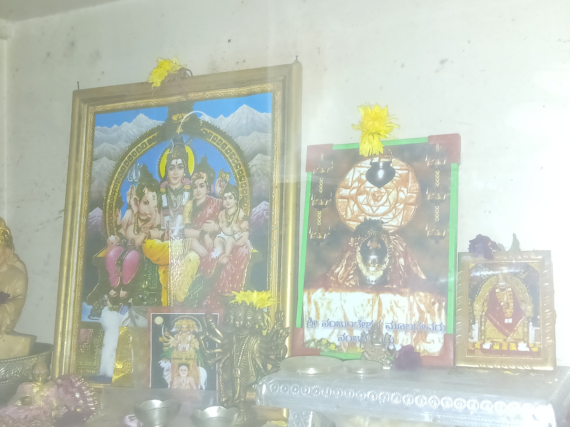 The image depicts a Hindu shrine, likely in a home in Bengaluru, India. The central focus is a shelf adorned with multiple framed pictures of deities, including Shiva and family, and other framed depictions. Fresh yellow flowers are placed atop several of the framed images. Small metal bowls, likely used for offerings or rituals, are positioned in the foreground. A brass statue of a multi-armed deity stands to the left of the bowls. A statue of a seated deity is also present to the left of the central picture. The backdrop is a plain white wall, indicating an indoor setting.