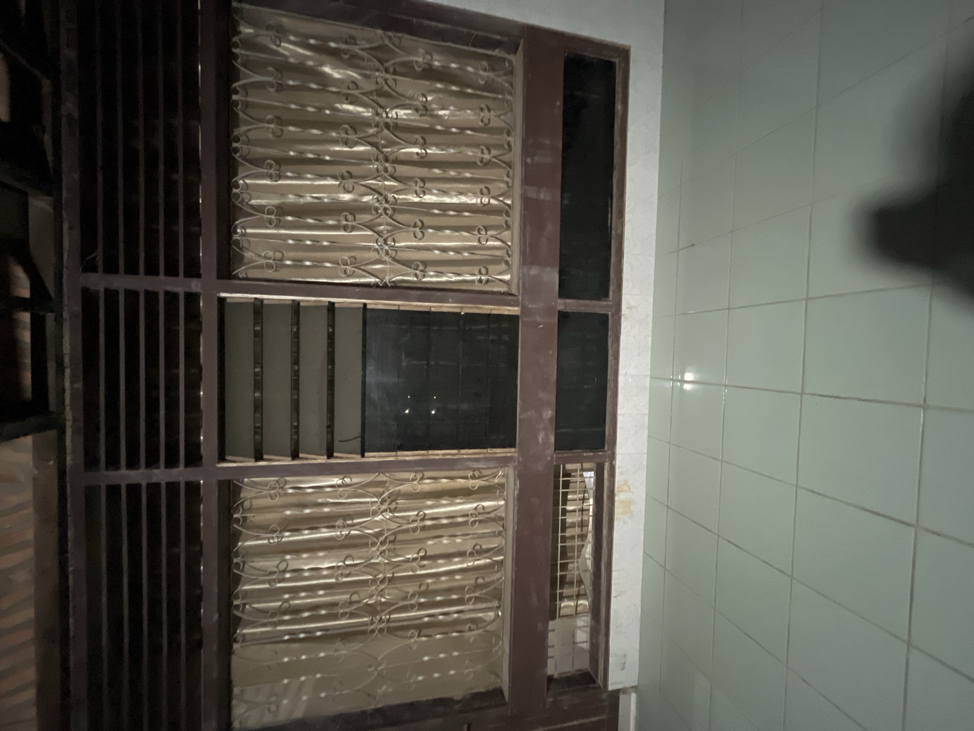 The image depicts a close-up of a window with decorative metal grates over a cream-colored curtain. The window is framed by dark brown wood. Below the main window, there is a lower section with a dark, reflective surface. The floor in front of the window is tiled with light green square tiles, and a shadow of a person is visible on the floor. The background shows wooden beams and a dark ceiling. The scene is likely an interior residential or commercial space.