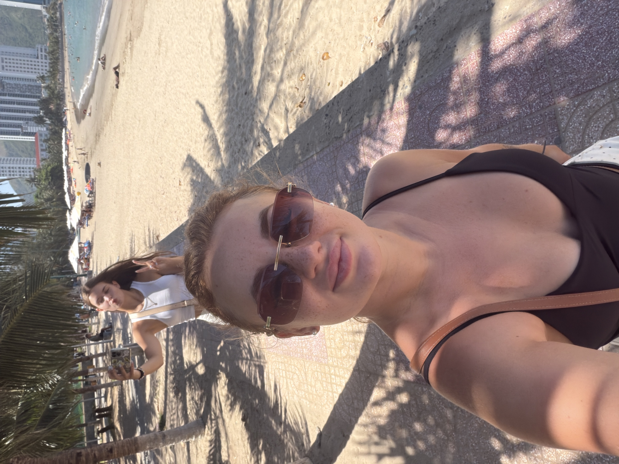Two young women are on a paved walkway next to a sandy beach in Nha Trang, Vietnam. The woman in the foreground is taking a selfie, with her face and upper body filling most of the frame. She is wearing a dark tank top and sunglasses. Her hair is pulled back, and she has freckles on her face. The woman in the background is also taking a selfie with her phone and making a peace sign with her fingers. She is wearing a white tank top and has her hair down. In the background, there is a beach with people sunbathing, umbrellas, lounge chairs, and the ocean. Tall buildings are visible in the distance. Palm trees line the walkway and the beach. The sun is shining, casting shadows on the walkway.
