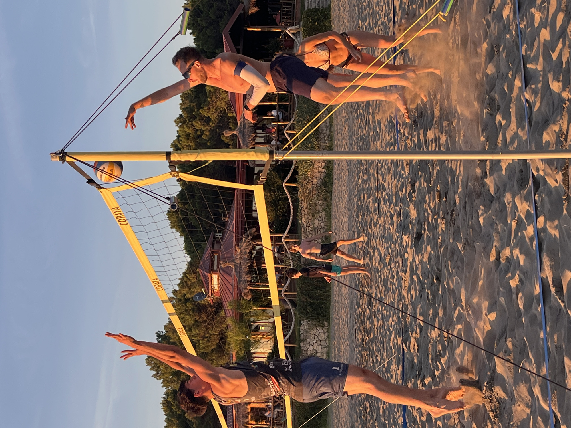 In the city of Pistula, Montenegro, four individuals are playing beach volleyball on a sandy court during what appears to be late afternoon or early evening, as indicated by the warm, golden light. Two men are actively engaged in the game on one side of the net, with one in the foreground leaping to hit the ball, his body angled towards the net, while his teammate stands behind him. On the other side of the net, a man is in the process of serving or hitting the ball, and a woman in a bikini is positioned behind him, observing the play. The beach volleyball net is strung between two yellow poles, with the word "COPAYA" visible on the net. In the background, trees and buildings with thatched roofs suggest a resort or restaurant area, adding to the coastal atmosphere. The sand is disturbed in several areas, particularly around the players' feet, showing the dynamic nature of the sport.
