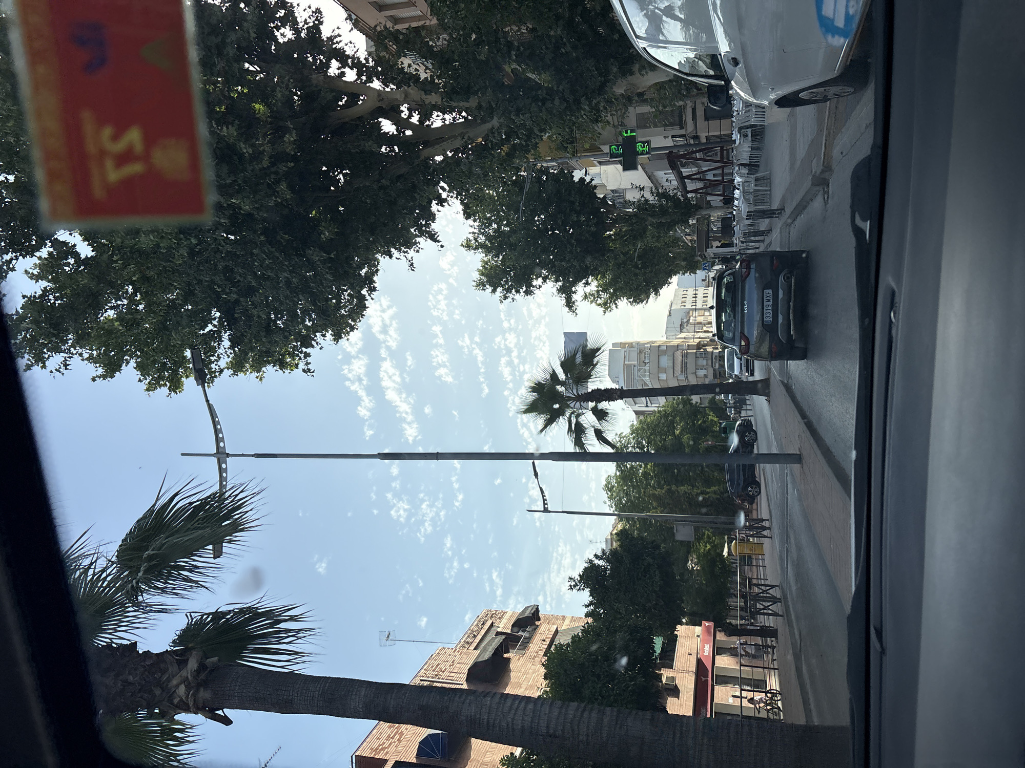 The media file depicts a street scene from the perspective of a vehicle, likely in Puente Genil, Spain, based on the provided context. The time of day appears to be daytime, with bright sunlight casting shadows. The sky is a clear blue with scattered white clouds, suggesting fair weather.
Prominently featured are several trees, including palm trees with their distinctive fronds, and deciduous trees with dense green foliage. Buildings line the street, appearing to be a mix of residential and commercial structures, some with visible architectural details and balconies.
Several vehicles are present on the road, including a dark-colored Kia with a license plate "B319 MXF". There are also parked cars and a white vehicle. Along the sidewalk, numerous empty white chairs and tables are set up, hinting at an outdoor dining area for a restaurant or cafe, which is currently not in use. A traffic light with green lights is visible in the distance.
There is a red sticker on the windshield of the vehicle from which the photo is taken. The sticker is partially obscured but shows some text and symbols, including what appears to be "S7".
The scene is characterized by its urban setting, the presence of nature in the form of trees, and the everyday activities of a street with vehicles and establishments. The overall mood is peaceful and sunny.
