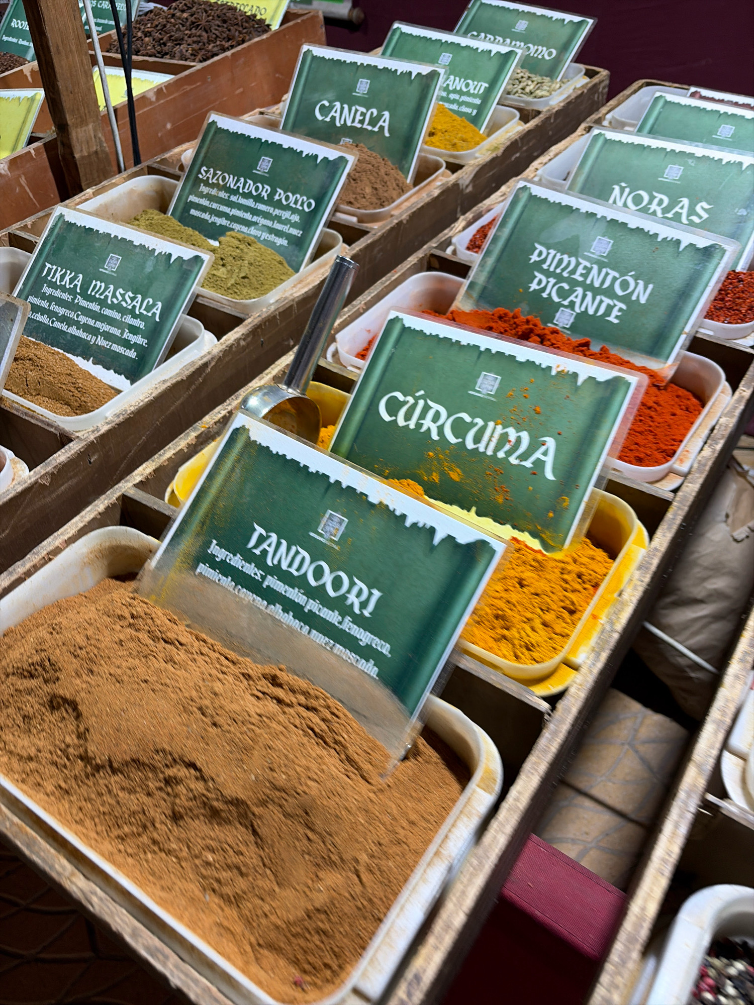 This is a close-up, eye-level photograph of a market stall in Salou, Spain, displaying various spices. The foreground is dominated by a large bin filled with finely ground reddish-brown spice, likely cinnamon or a spice blend. Several smaller bins, each with a green label indicating the spice's name, are visible behind and to the sides.
The labels read:
* "TIKKA MASSALA" with ingredients listed below.
* "SAZONADOR POLLO" (Chicken Seasoning) with ingredients listed.
* "CANELA" (Cinnamon).
* "CURCUMA" (Turmeric).
* "NORAS".
* "PIMENTÓN PICANTE" (Hot Paprika).
Some of the bins contain the spices themselves, such as the vibrant yellow turmeric and the deep red paprika. A metal scoop rests in the bin of turmeric. The stall appears to be made of rustic wooden crates, typical of market displays. The background is slightly blurred, showing more bins of spices and the general market environment. There are no people or animals visible in this frame.