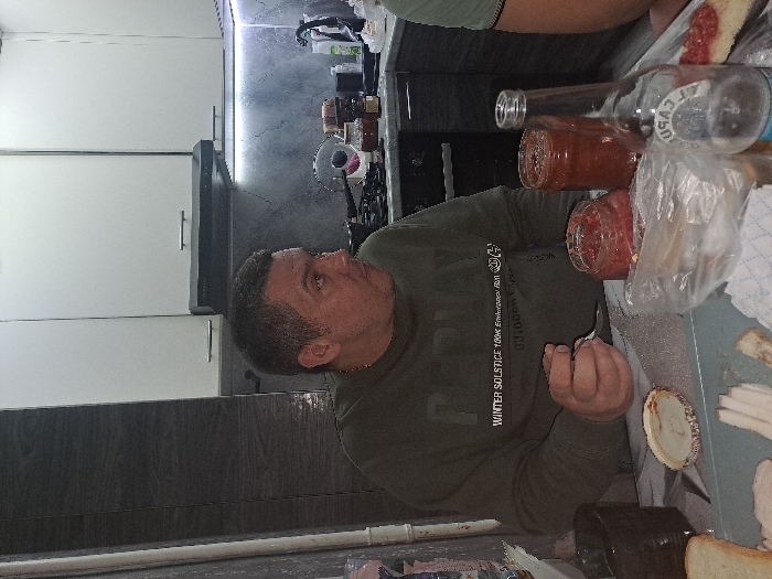 A man wearing a green sweater sits at a table laden with food. There are jars of tomato sauce, a clear glass bottle, and a plastic bag. The table holds bread, what appears to be pieces of fat, and other unidentifiable items. The background shows white cabinets and a dark backsplash. The image has been rotated counterclockwise.