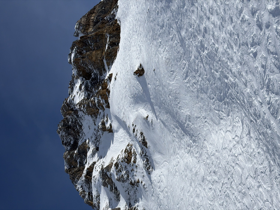 An image captured in Courchevel, France, depicts a steep, snow-covered mountain face under a clear, deep blue sky. The upper third of the frame reveals rugged, rocky peaks and ridges, predominantly dark brown and grey, partially covered with white snow. Sunlight illuminates these formations, creating contrasts between the rock and snow. The lower two-thirds of the image present a wide, expansive snowfield, heavily marked with numerous intersecting ski tracks. These tracks vary in width and depth, indicating multiple descents and turns across the slope. Several rocky outcrops protrude from the snow on the lower and mid-slopes, adding texture to the environment. No individuals, man-made structures, or equipment are visible within the frame, with human activity evidenced solely by the extensive ski tracks.
