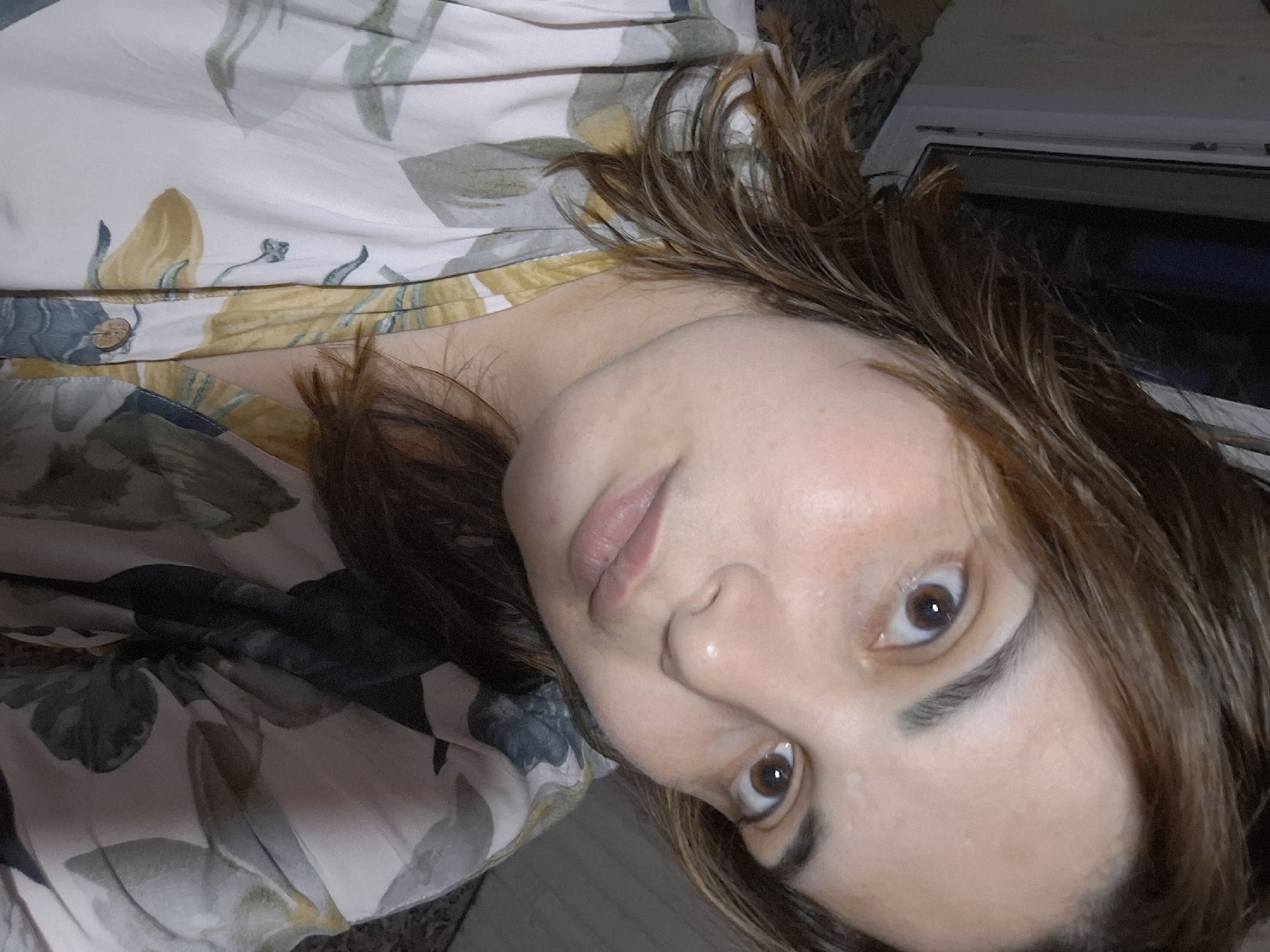 A close-up, slightly angled headshot photograph shows a fair-skinned woman with brown hair and brown eyes. She is wearing a floral print shirt with a yellow and grey design. Part of a window is visible in the background. There is no indication of the location being in the city of Амзя, Russia.