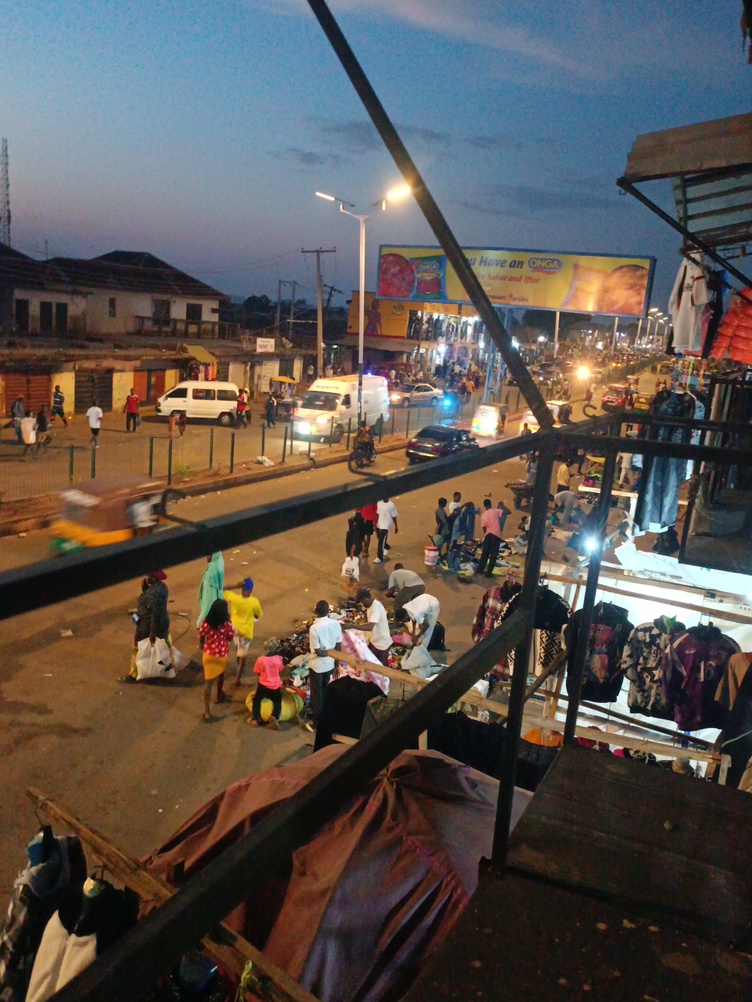 This image shows an evening street scene in Bukuru, Nigeria. The photograph is taken from an elevated position, possibly from a building or structure overlooking the street. A variety of people are present, including pedestrians and vendors. Some people appear to be shopping or browsing through items displayed on the ground. There is a noticeable accumulation of goods or merchandise spread out on the ground, indicating a makeshift market or informal trading area. Several vehicles are visible on the road, including cars, an ambulance, and a van. A large billboard advertising "ONGA" seasoning stands in the background. The lighting suggests that it is dusk or early evening, with streetlights illuminating the scene. There are metal structures and clothing racks visible in the foreground. The sky is a dusky blue with scattered clouds.