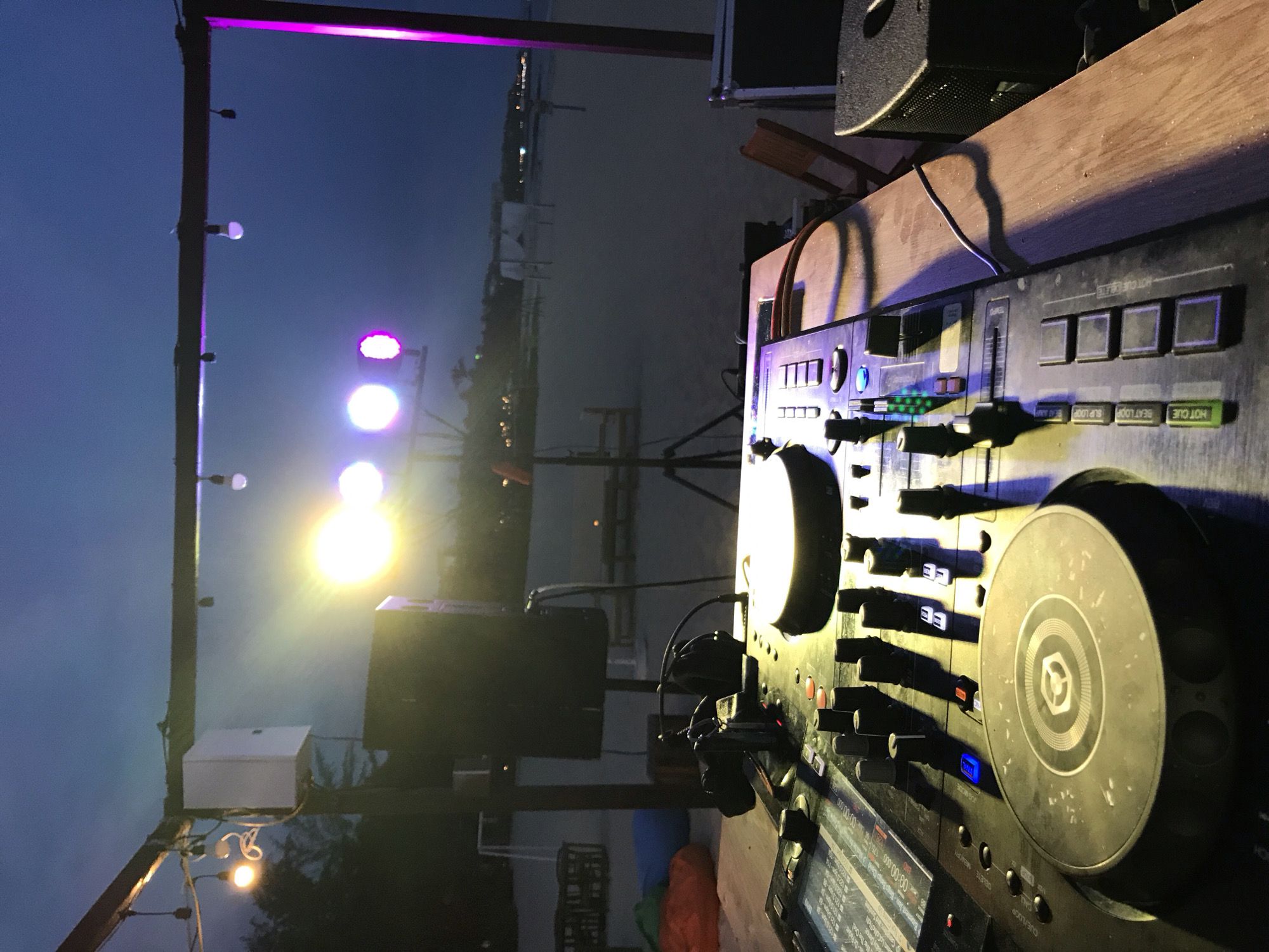 A DJ console is set up on a wooden table on a beach at dusk. The console has several buttons, knobs, and two turntables. In the background, there are beach chairs, umbrellas, and the ocean. Lights are illuminating the scene, with some purple and yellow lights visible. The sky is a dark blue.