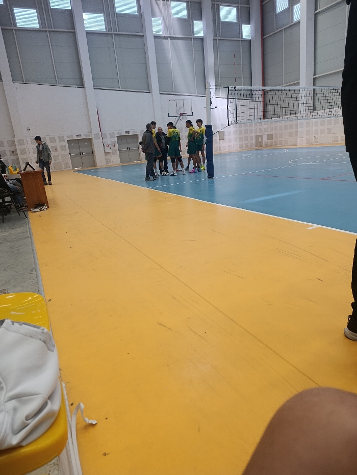 An indoor sports hall in Zavet, Bulgaria, is depicted, featuring a multi-sport court. The foreground floor is yellow, transitioning to a light blue playing area marked with white lines. A volleyball net, black with a white top band, is set up across the blue court. A basketball backboard and hoop are visible on the far white wall behind the volleyball net.
Approximately eight to ten individuals are clustered in a huddle on the blue court, positioned near the volleyball net. Most of these individuals are young males dressed in athletic uniforms predominantly green and yellow, with numbers visible on some jerseys (e.g., "22"). One or two older males, dressed in darker, casual clothing, are also part of this group, possibly in coaching roles.
On the left side of the frame, near the yellow floor, a male in a light-colored jacket and dark pants stands next to a wooden table. Another individual is partially visible, seated behind this table. In the lower-left corner, a white garment is loosely placed on a yellow plastic chair. The lower right portion of the image shows the lower leg and foot of a person wearing a black athletic shoe with a white sole, consistent with the photographer's immediate vantage point. The facility walls are white, with horizontal rectangular windows or vents positioned high along the ceiling, allowing natural light into the hall.