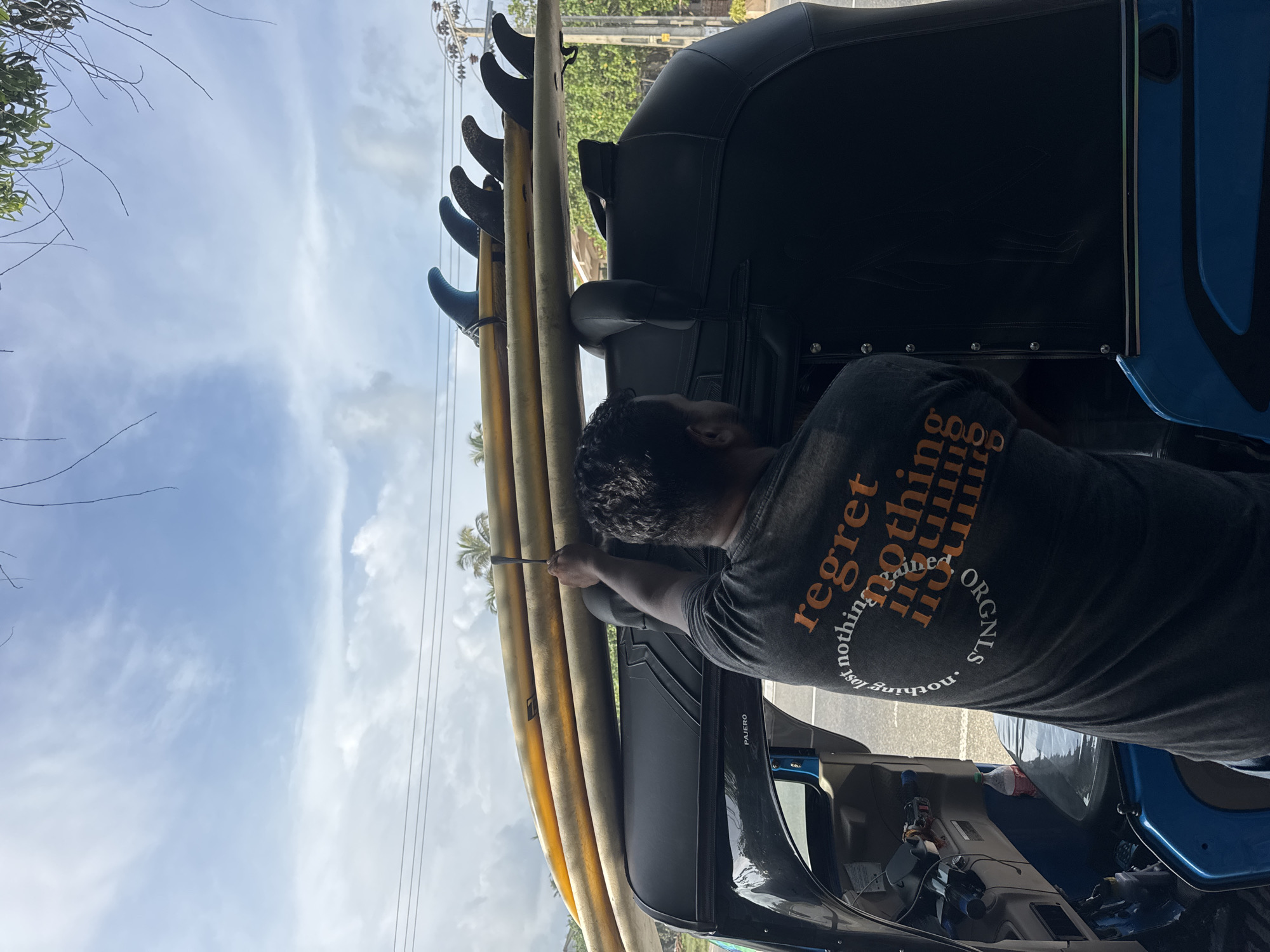 A man is securing surfboards on top of a blue vehicle in Kathaluwa, Sri Lanka. The boards are stacked vertically, with the fins pointing upwards. The man is using a tool to tighten the straps. The vehicle has a black soft top and is parked on a paved road. The sky is cloudy.