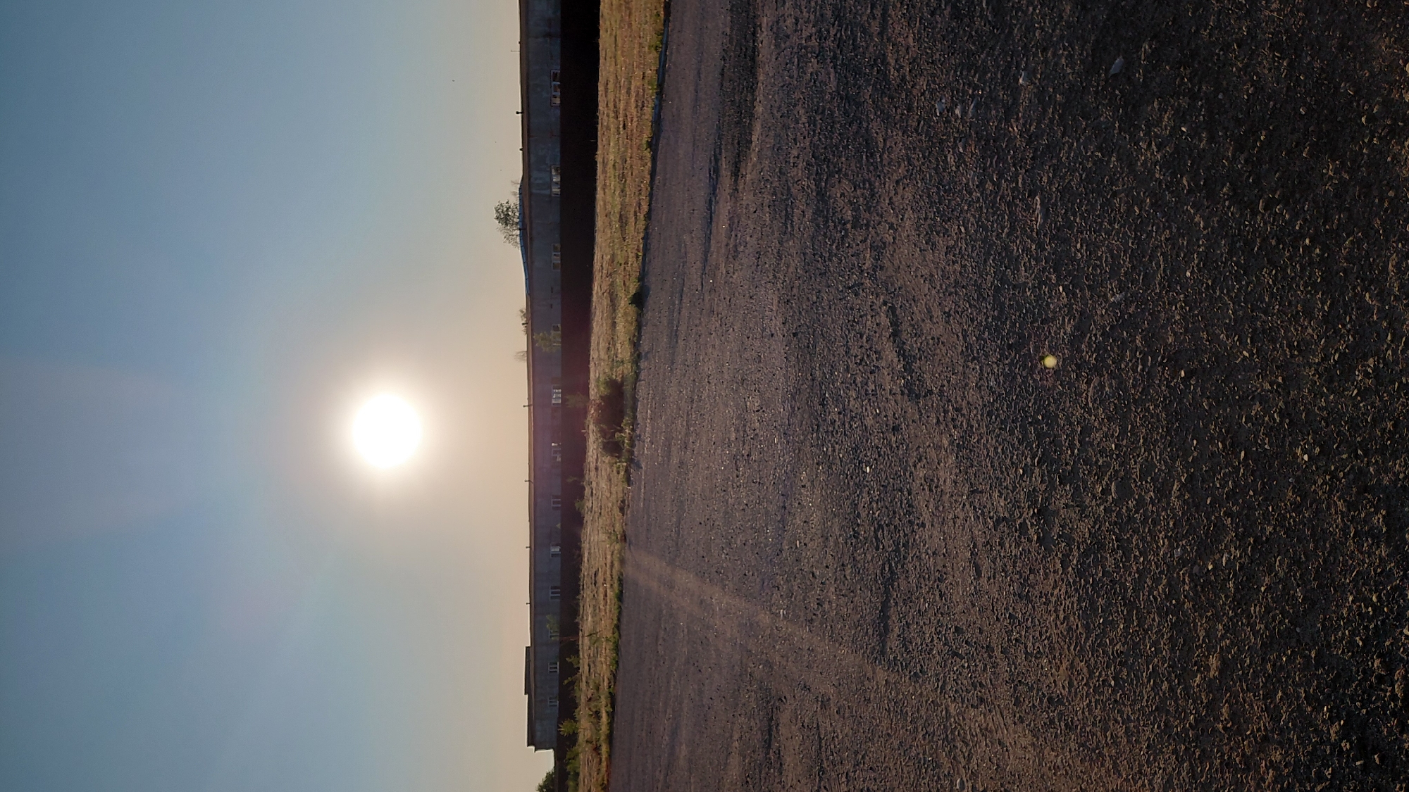 A photograph taken during the daytime shows a sun-drenched building and a dirt road. The sun is in the upper portion of the image, with light rays emanating from it. A long, low building with windows is visible in the middle ground, behind a dark fence. The area in front of the building is covered in dry grass and bushes. The foreground is a dirt road, with pebbles and tire tracks. There are no people visible in the image. The scene appears to be in an industrial or rural area, likely on the outskirts of a city. Based on the available information about the location of Saratov, Russia, this image could depict an area near an industrial zone or a less developed part of the city, characterized by open spaces and functional, utilitarian structures rather than residential buildings or significant greenery.