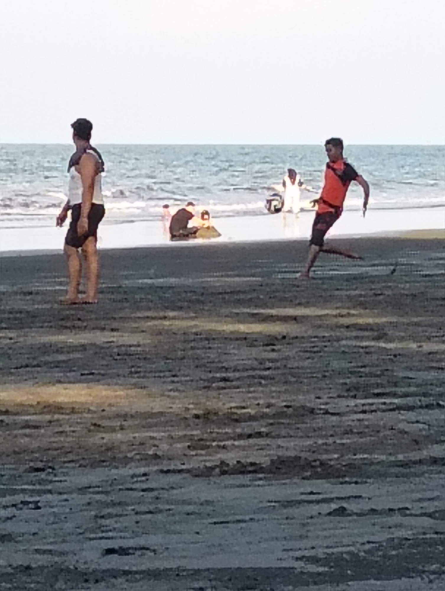 The image depicts a beach scene with several individuals. In the foreground, a person wearing a white tank top and dark shorts stands facing away from the camera. To the right, another person in an orange and black t-shirt and patterned shorts is captured in mid-stride, appearing to be playing soccer. A soccer ball is visible near their feet. In the background, near the water's edge, a group of people are gathered, some appearing to be sitting or kneeling. The ocean stretches out behind them under a bright sky. The beach itself is composed of dark sand. The overall setting suggests a recreational activity taking place at a coastal location in Peukan Sot, Indonesia.
