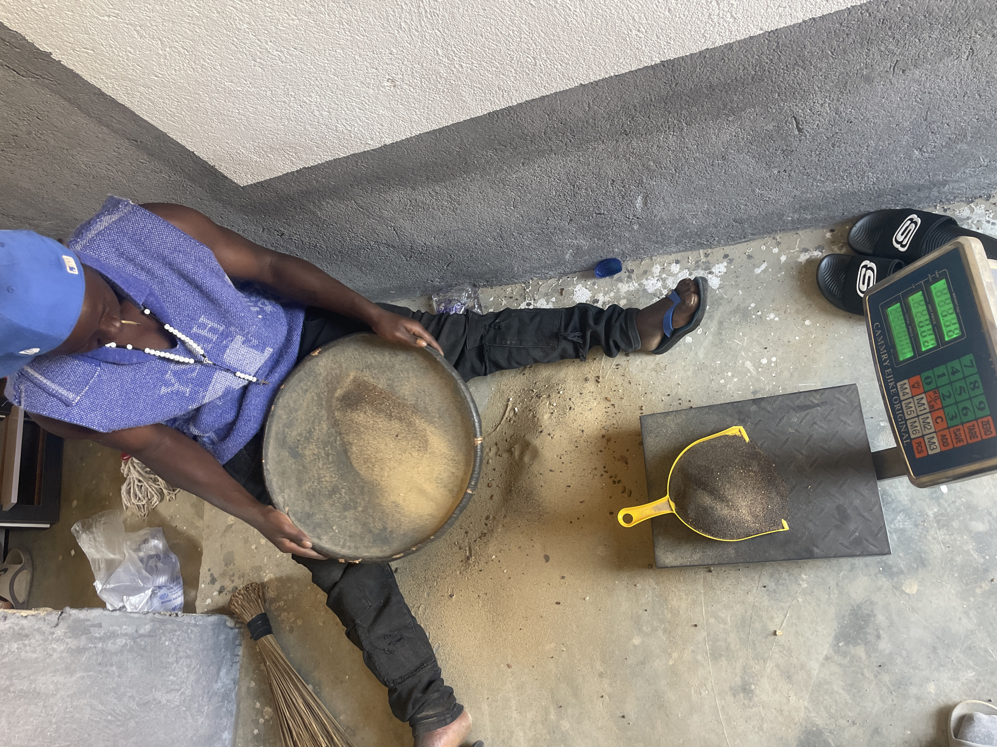 A person wearing a blue cap and sleeveless patterned shirt sits on the ground, holding a large, round, shallow pan containing a granular substance. The person is weighing the granular substance using a digital scale with a yellow scoop, placed on a black platform. A pair of black and white sandals rest near the scale. The floor is concrete with stains. The setting appears to be indoors, against a rough concrete wall.