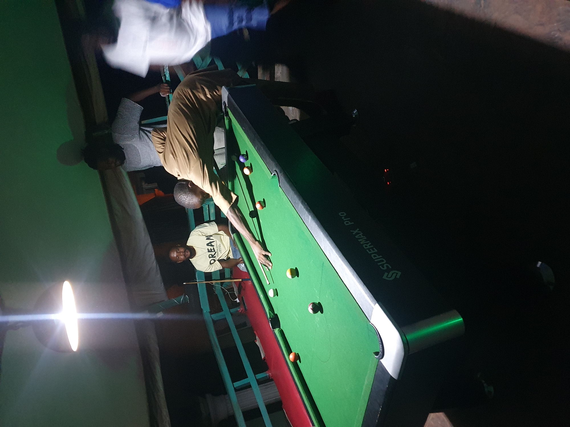 A group of men are playing pool in a dimly lit, outdoor setting, possibly on a balcony or patio, in Lokoja, Nigeria. A single, bright overhead light illuminates the pool table. One man, wearing a light-colored t-shirt with "DREAM" printed on it, sits watching the game. Another man in a brown shirt is actively playing, aiming a shot with a cue stick. Two other men are partially visible in the background. The pool table is green with several colored balls scattered across it. The environment appears somewhat rustic, with railings and a wooden ceiling visible.