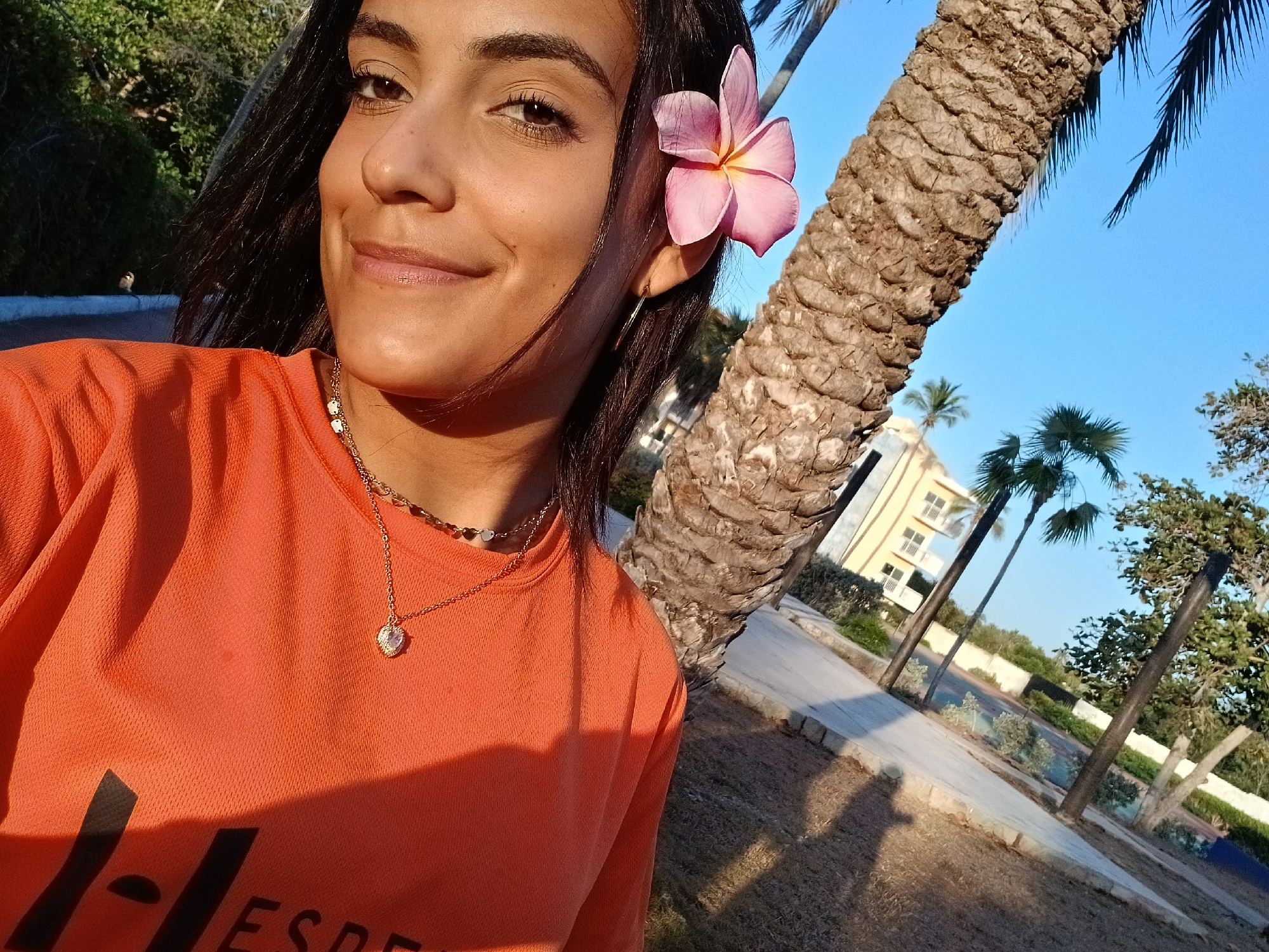A woman is taking a selfie outdoors, likely in Playa El Agua, Venezuela, given the tropical environment. She is wearing an orange t-shirt and a necklace, and has a pink flower tucked behind her ear. A palm tree trunk is visible to her right. In the background, there's a building and more palm trees under a clear blue sky. The lighting suggests it's either early morning or late afternoon.