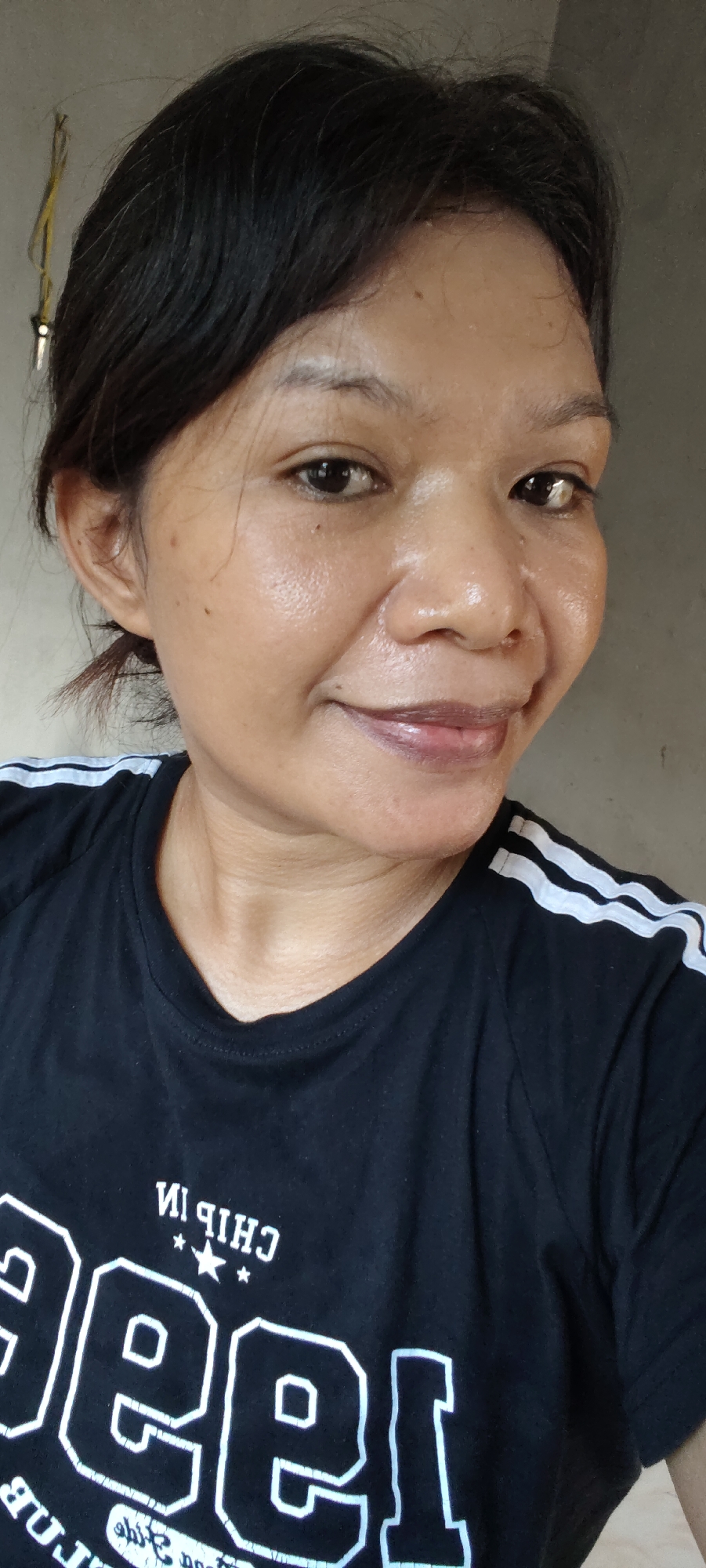 A medium close-up, head-and-shoulders shot shows a young woman with dark hair pulled back, wearing a black t-shirt with white stripes on the shoulders and white lettering. She has a light brown complexion and is looking slightly off-camera to her right, with a subtle smile. Her skin appears slightly oily. The background is a neutral, textured wall. This image was taken in Sika Makmur, Indonesia.