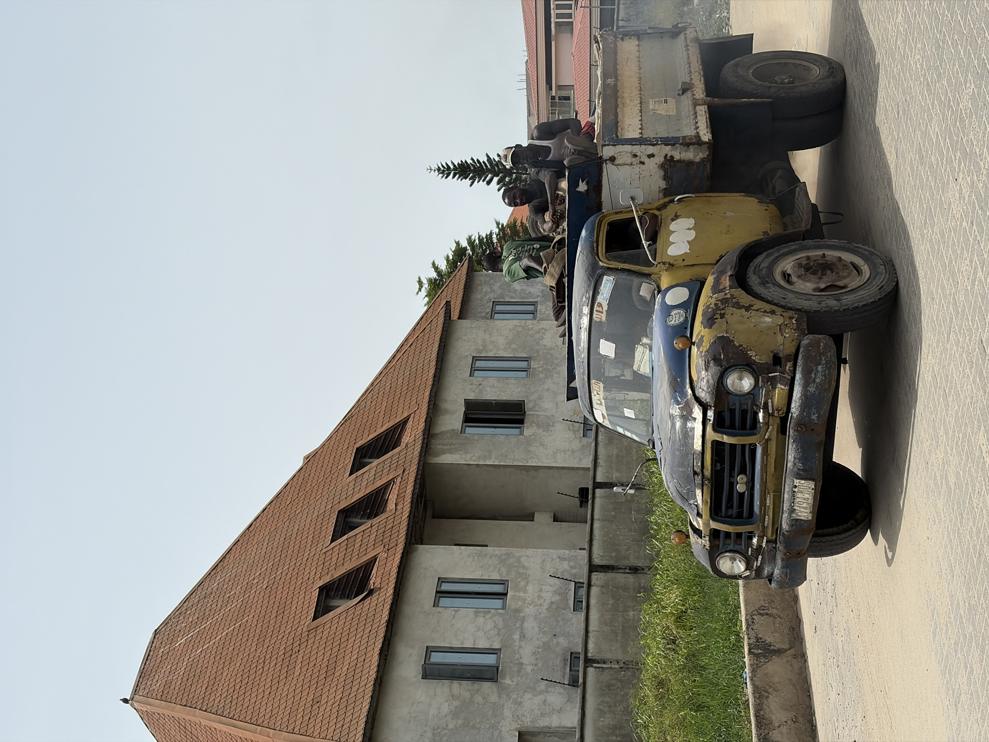 The image depicts a dilapidated, yellow and blue truck parked on a concrete surface in Igboefon, Nigeria. The truck's condition is poor, with visible rust and damage. Several men are riding in the back of the truck. Behind the truck is a building with a red-tiled roof and a gray facade featuring multiple windows and balconies. The sky is overcast. A pine tree is visible behind the building. A license plate on the front of the truck reads "LAGOS Y496 MUS".